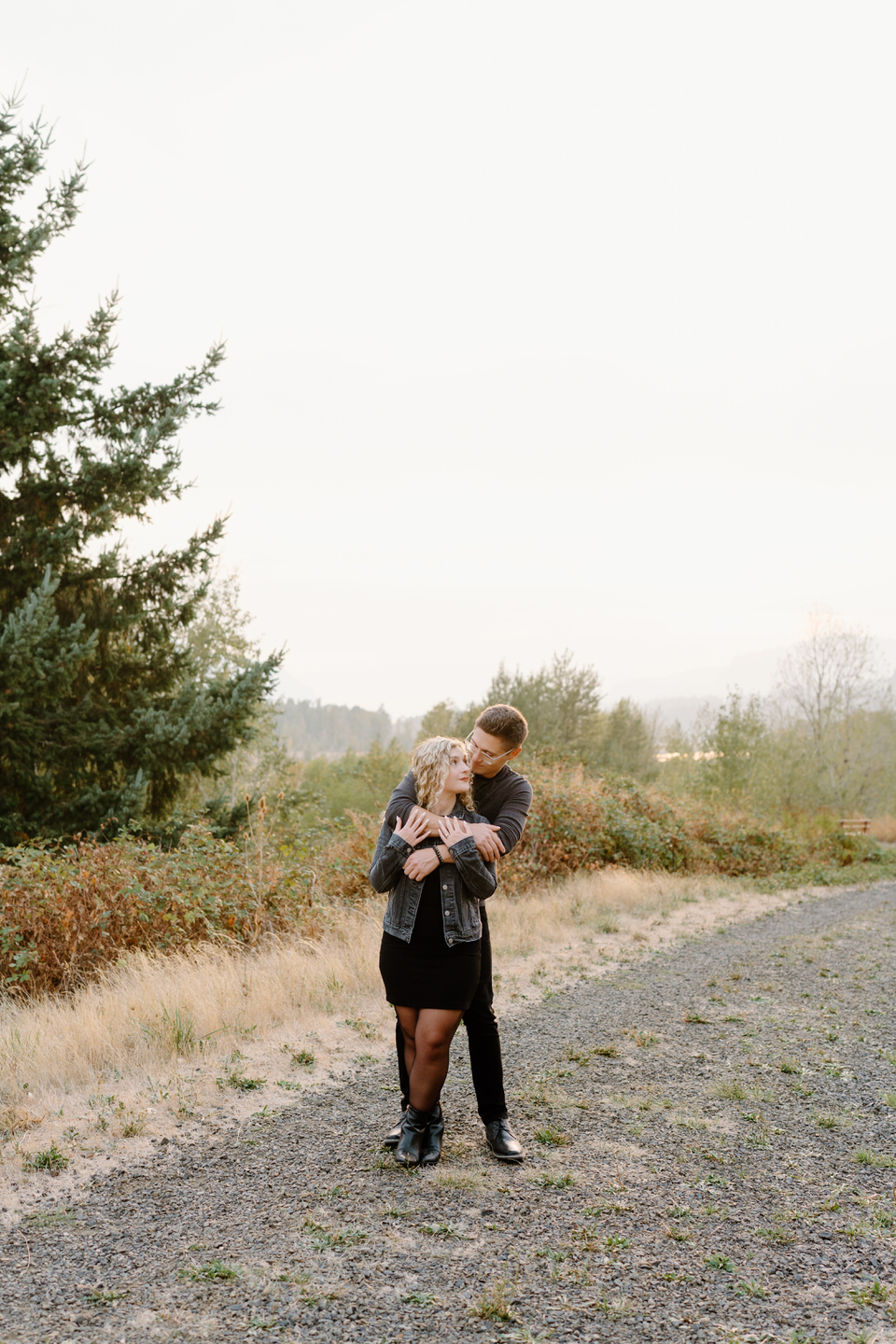Man wraps his arms around woman, both in dark outfits among the light background.