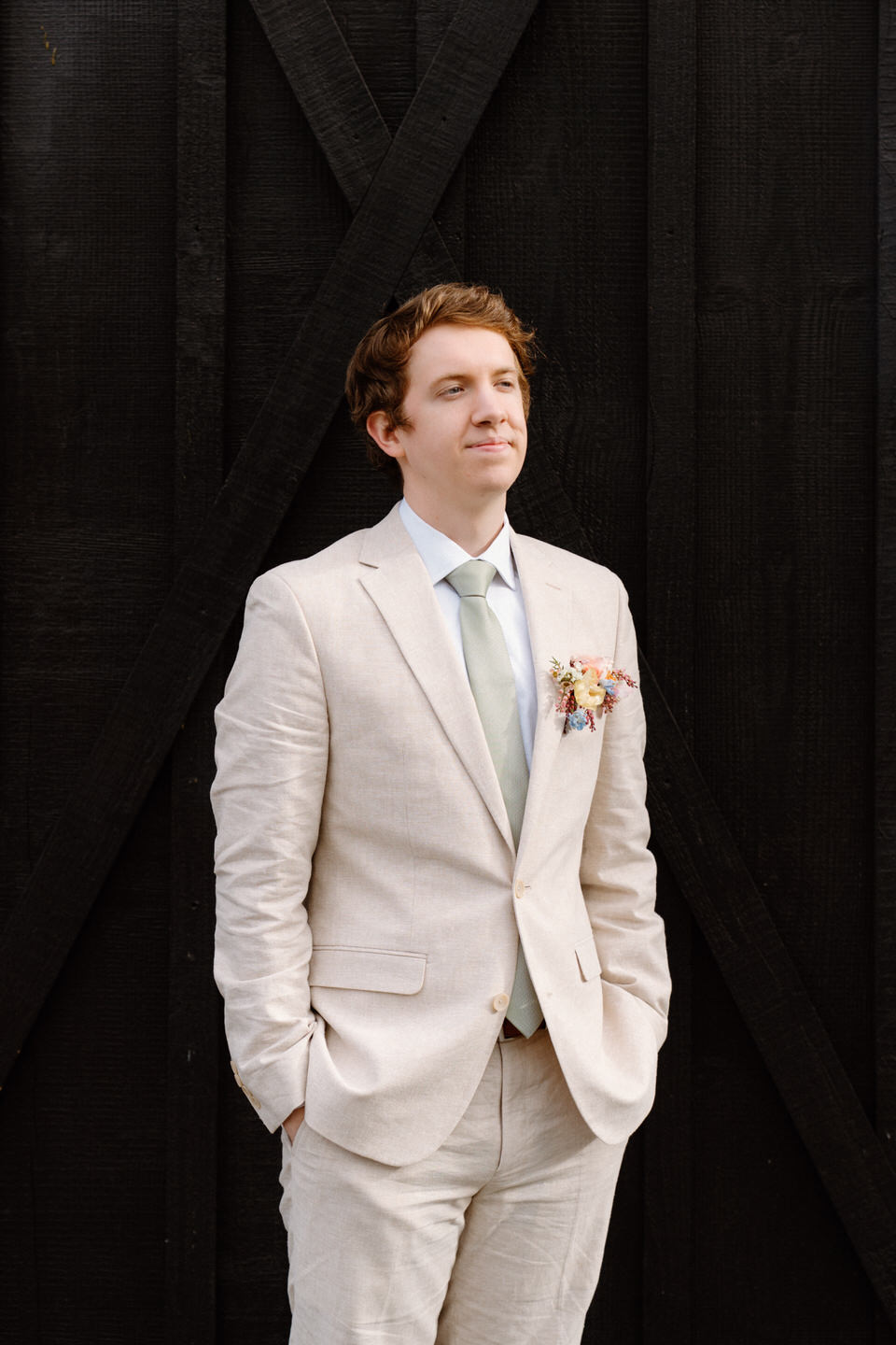 A tan suit contrasts beautifully against the dark wood barn exterior at Sage & Social’s reception space.
