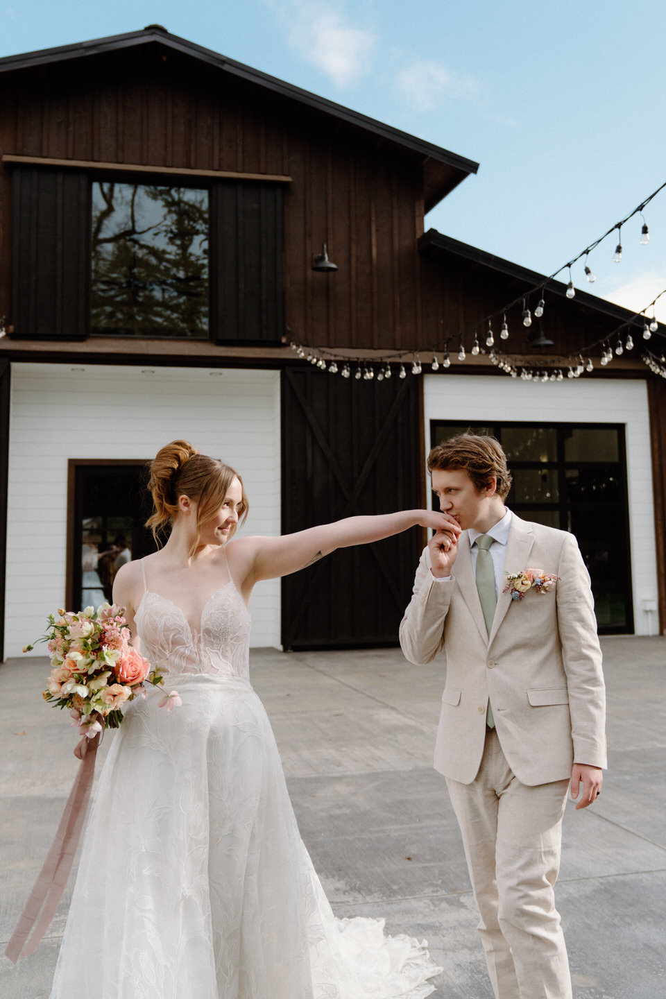 A couple shares a playful moment, walking in front of the barn at Sage & Social under soft spring light.