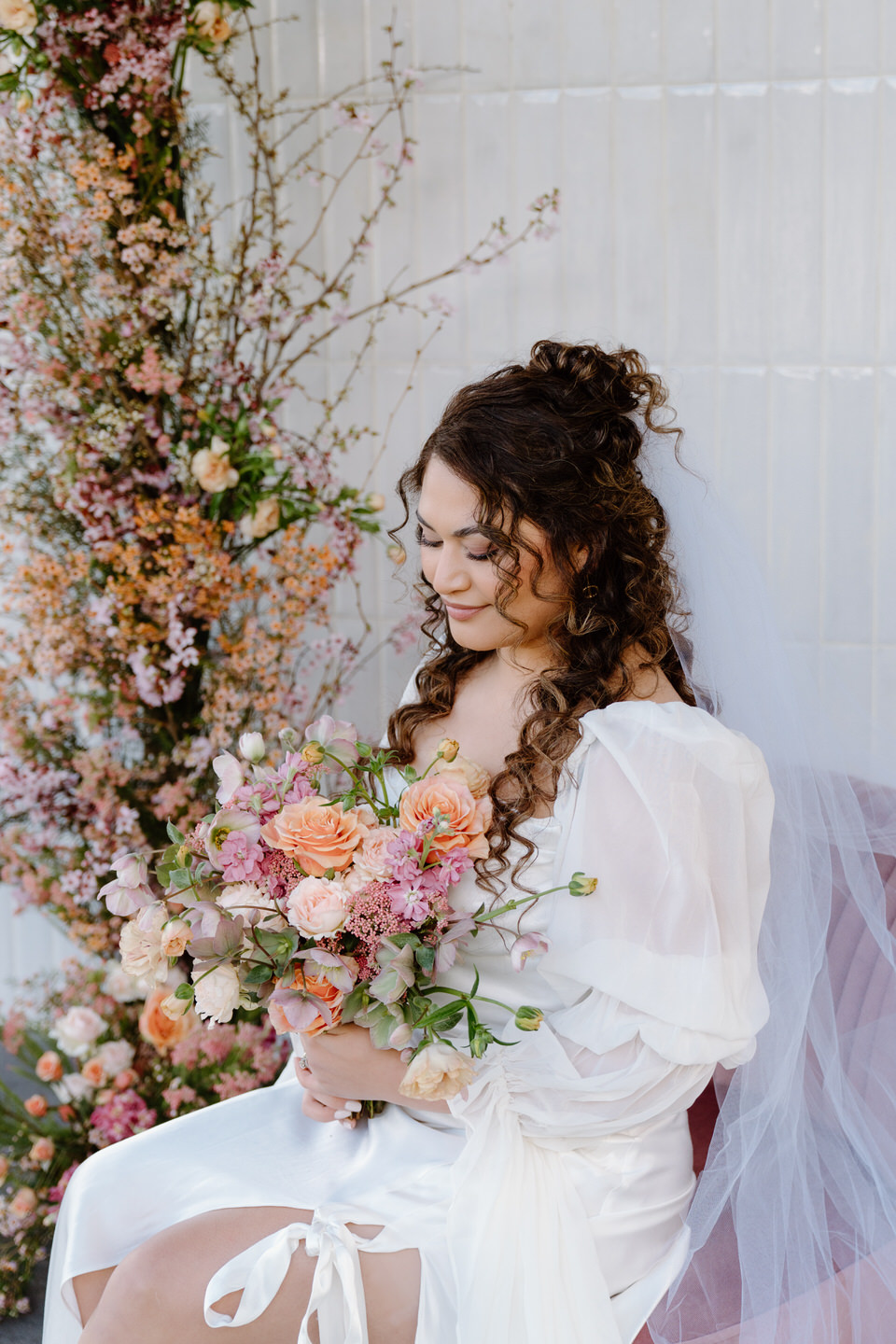 The bride smiles, seated on the pink chair with veil draped behind, glowing in the natural Sage & Social light.