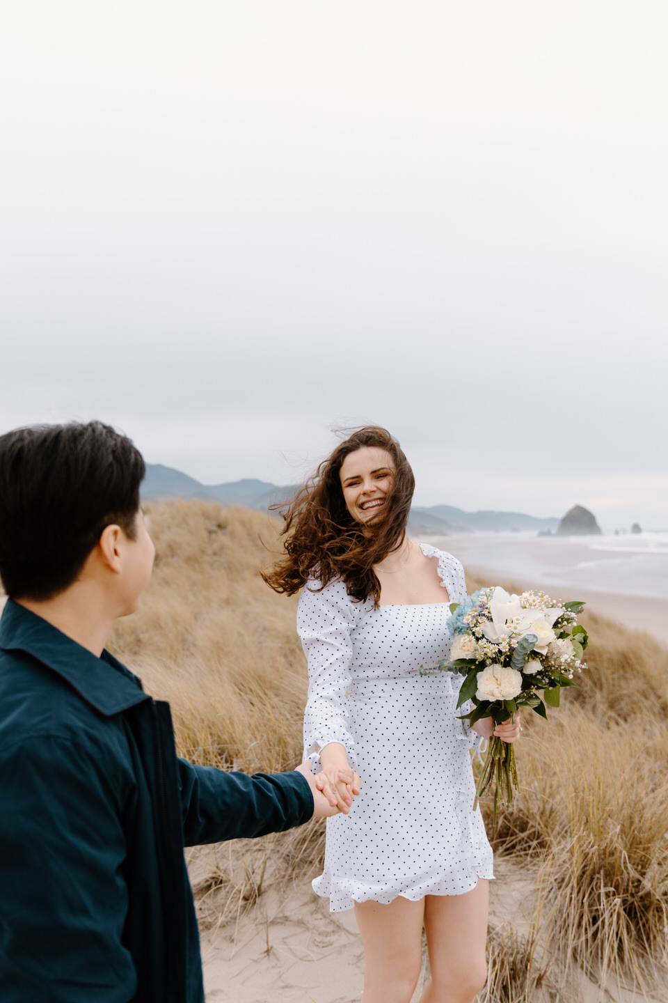 Wind blowing woman’s hair as couple walks through grassy dunes by the beach.