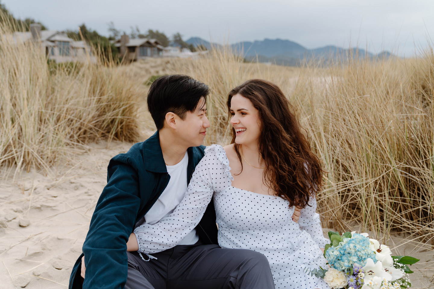 Windswept engagement photo with couple smiling by coastal dunes.