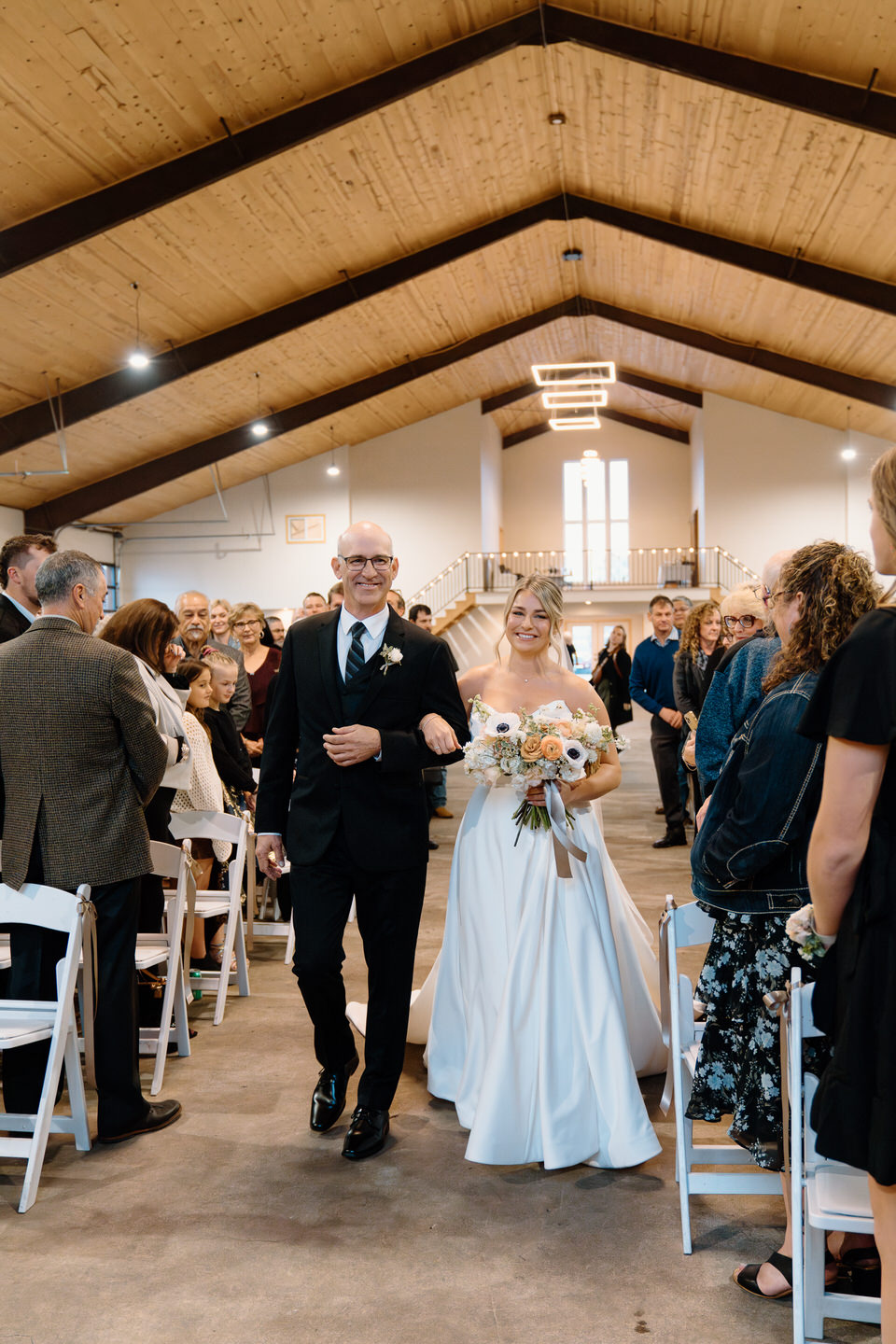 Bride and her father walk down the aisle in the indoor barn.