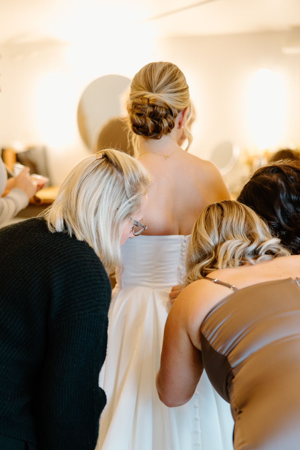 Bridesmaids and family members help bride button up her dress from the back. 