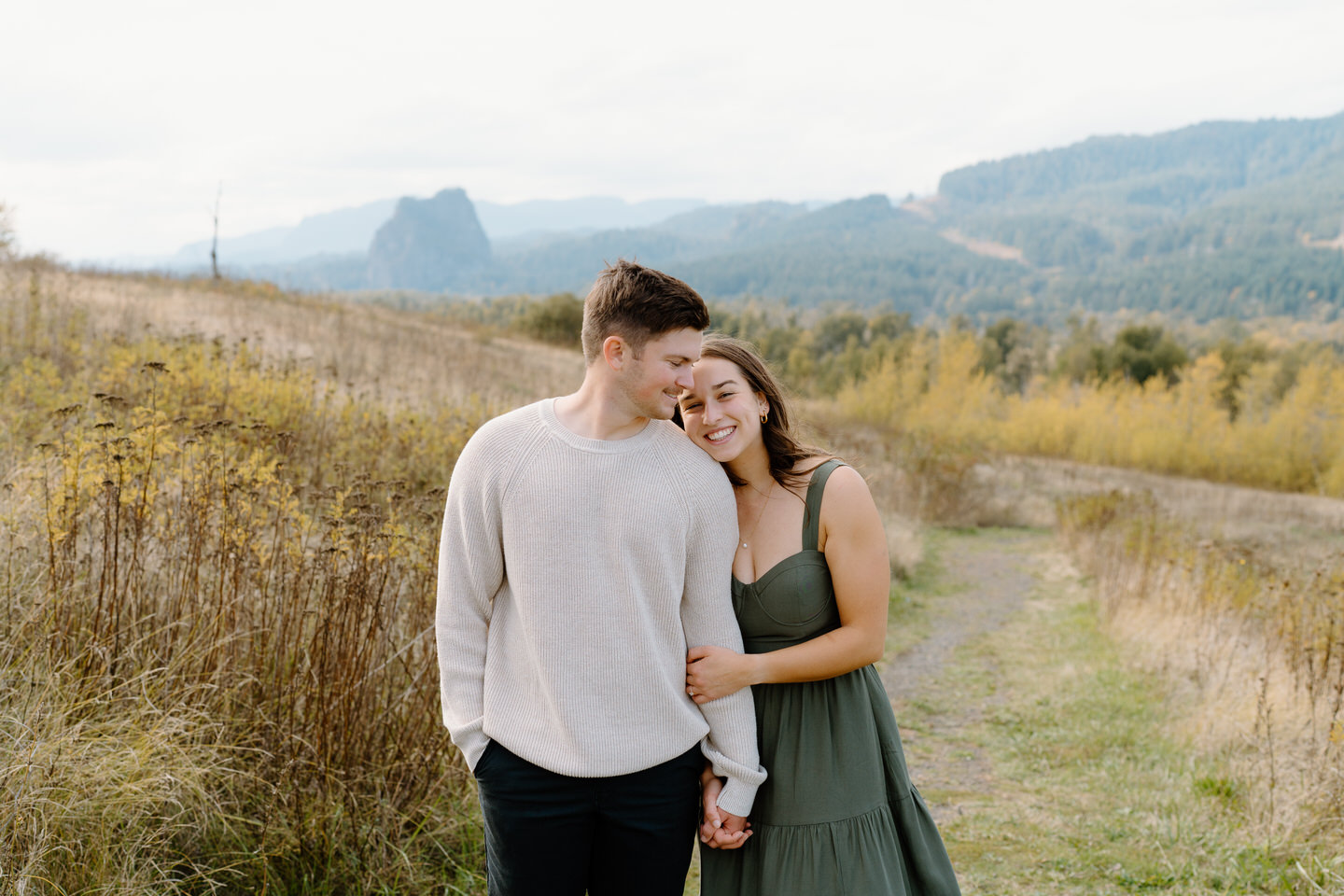 Smiling couple in green and brown outfits hugging under cloudy skies — fall engagement photo in Oregon’s Columbia Gorge.