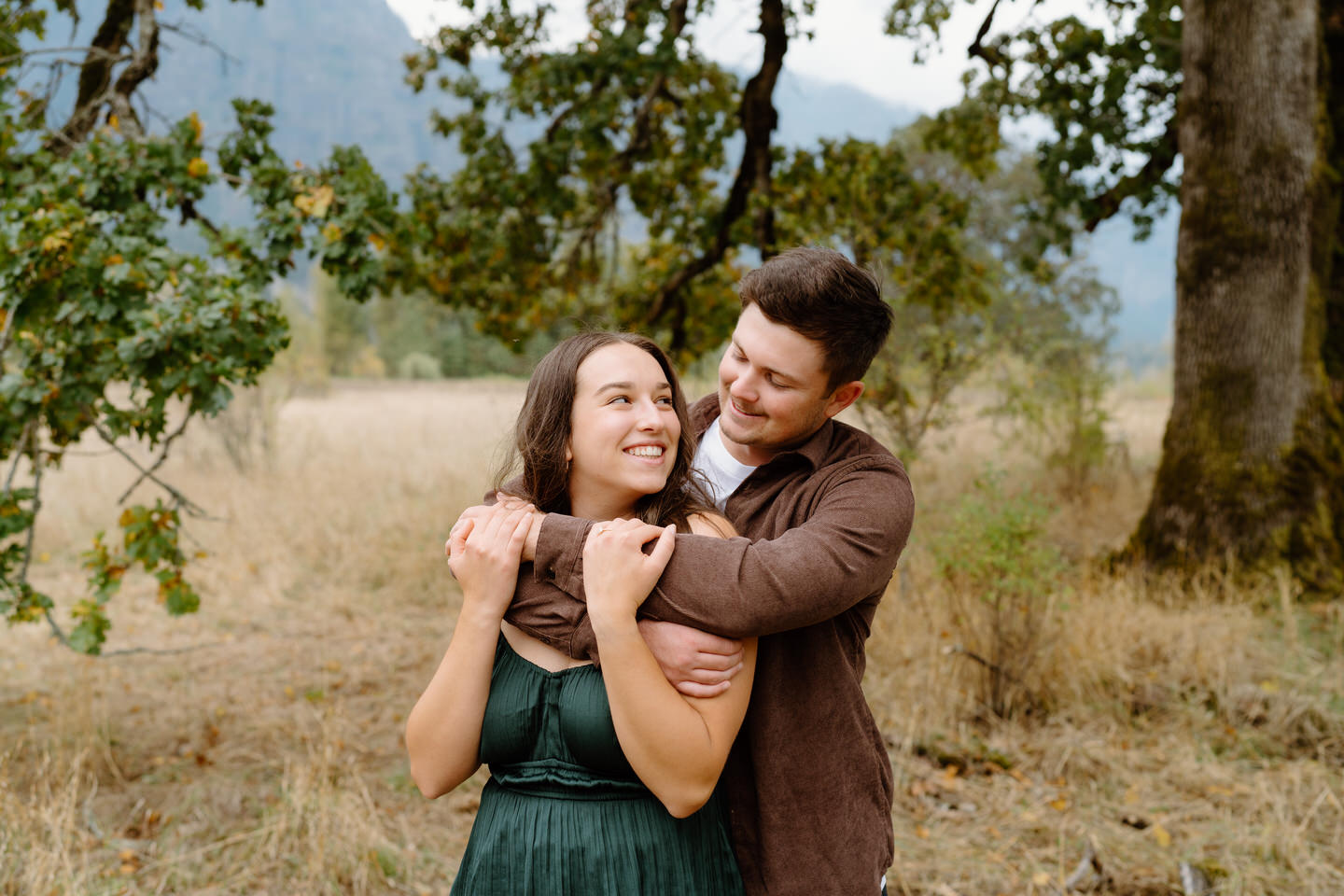 Cozy engagement photo with man hugging woman from behind, both laughing — fall couple photo idea.