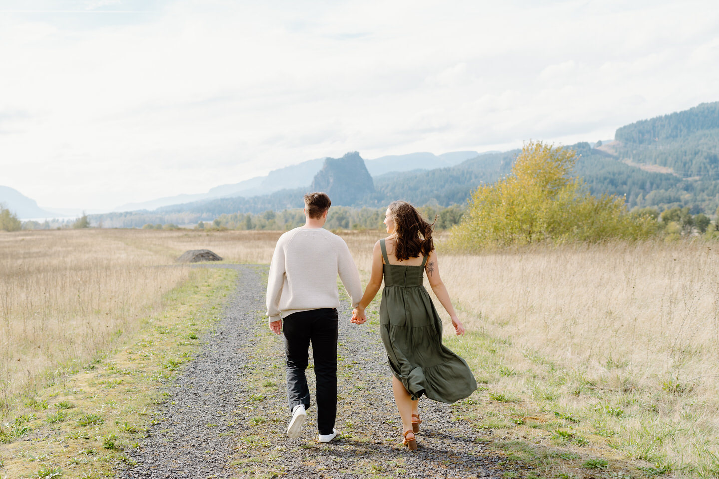 Full-body shot of engaged couple walking away on a dirt path, surrounded by golden fall grass.