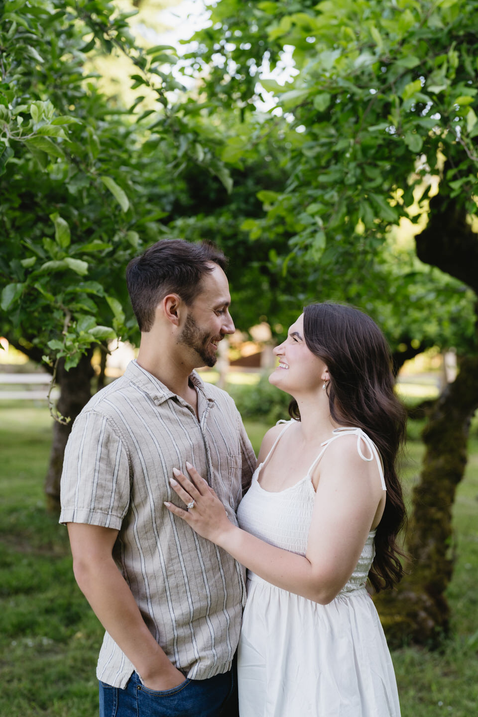 Couple stands in Sauvie Island orchard, her hand on his chest to show engagement ring.