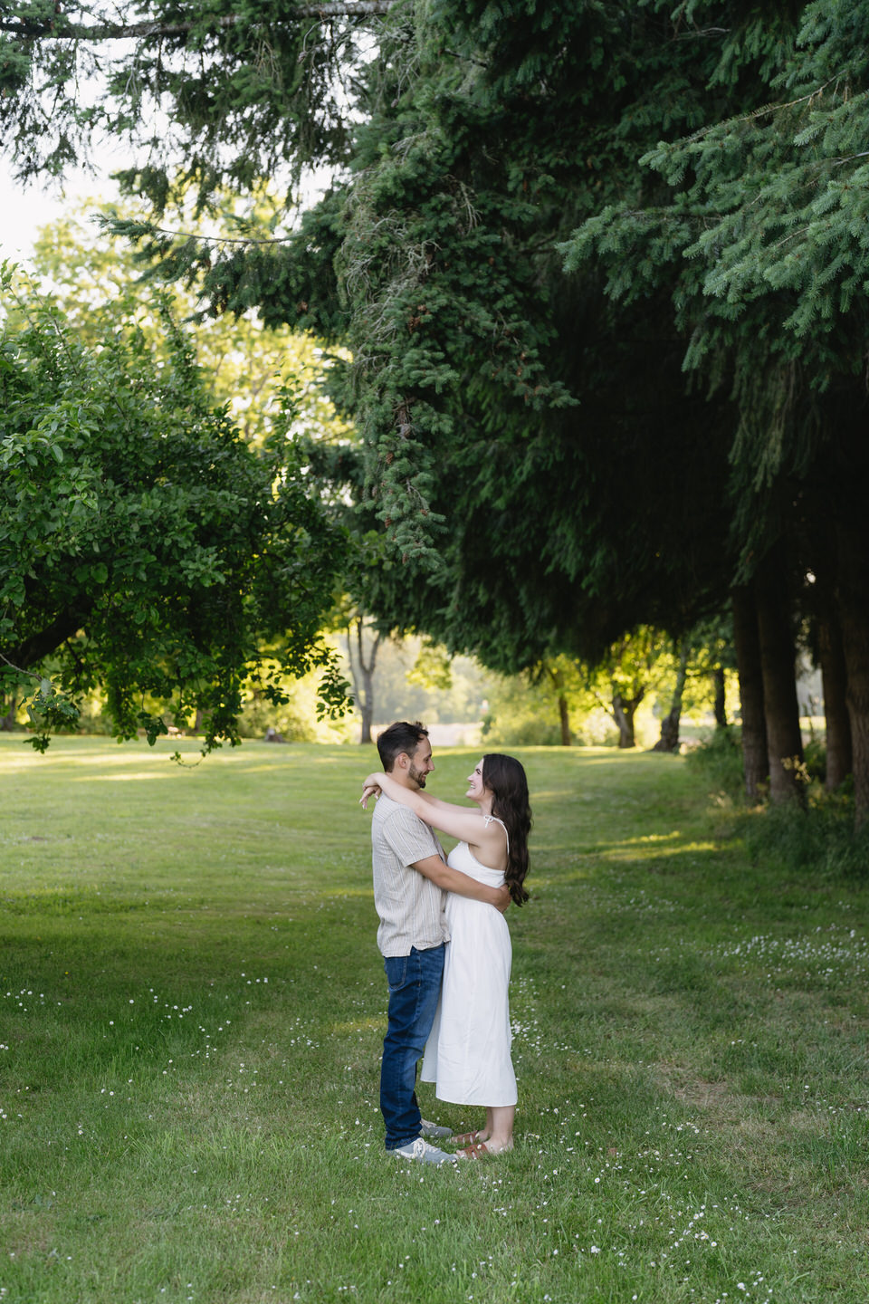 Bride and groom-to-be with arms wrapped around each other among evergreen trees.