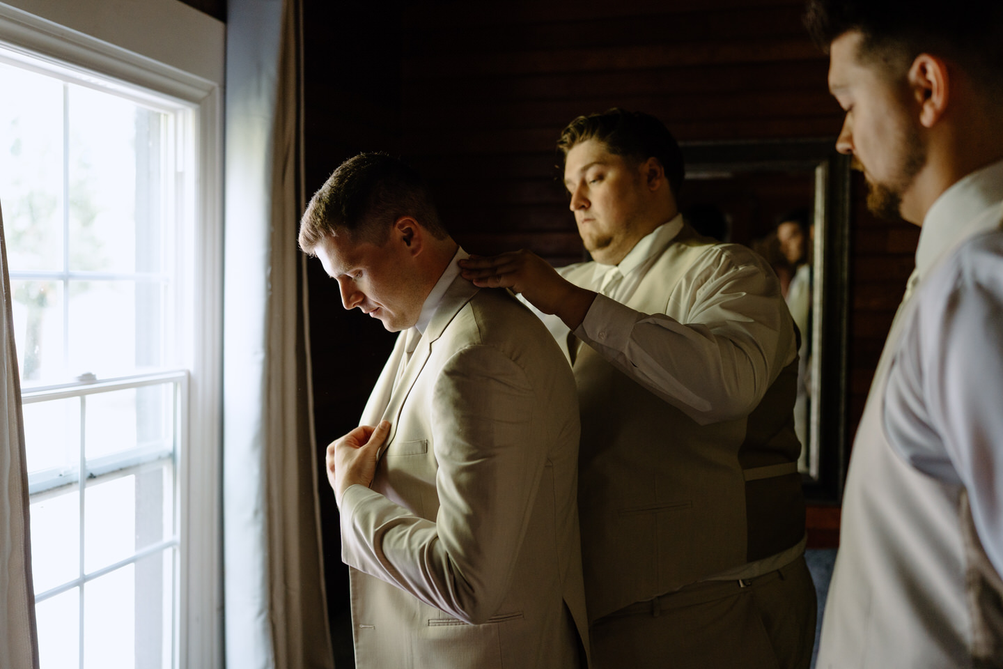 Groomsmen help the groom adjust his coat as he looks out the window of the morning suite.