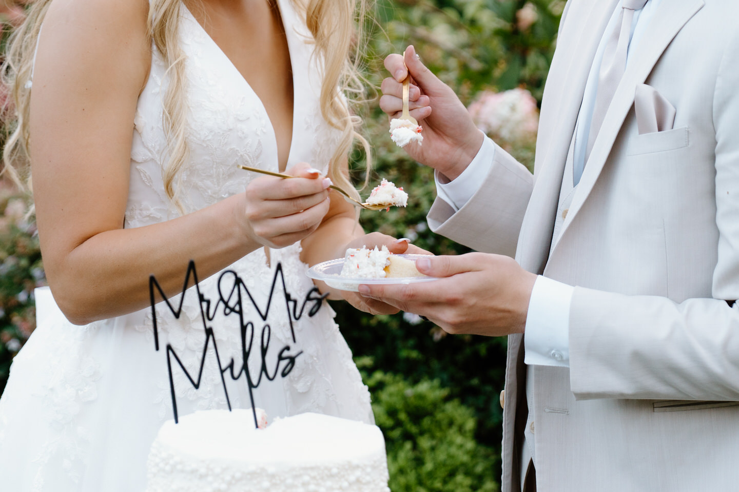 Close up of a couple sharing a slice of wedding cake with their "last name" cake topper in front of them.