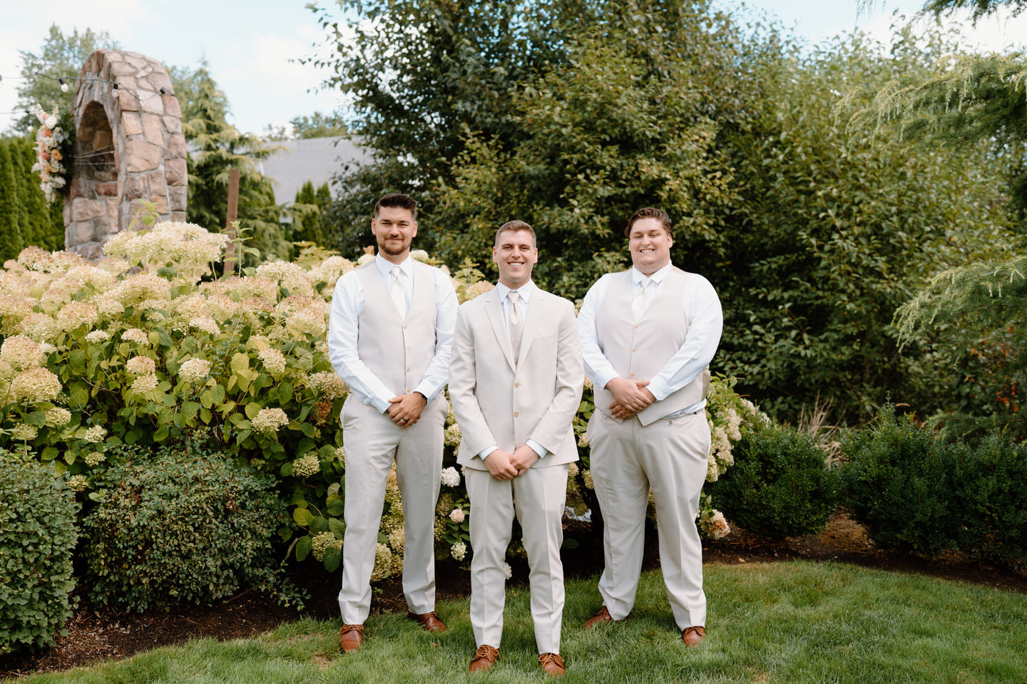 Groom stands in front of his groomsmen, smiling with their hands held in front of them.