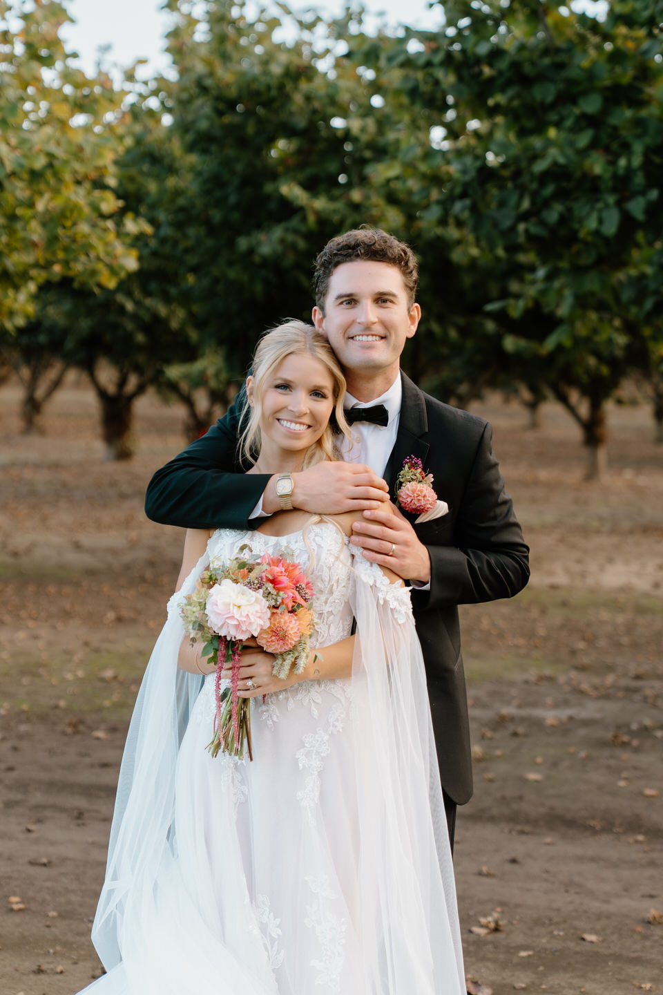 Bride and groom smile at camera while the groom wraps his arms around her.
