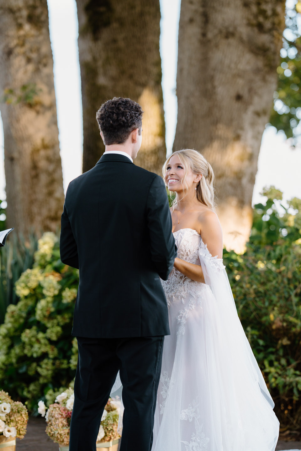 Bride smiles cheerfully at her groom, holding hands among the oak trees at Postlewait's.