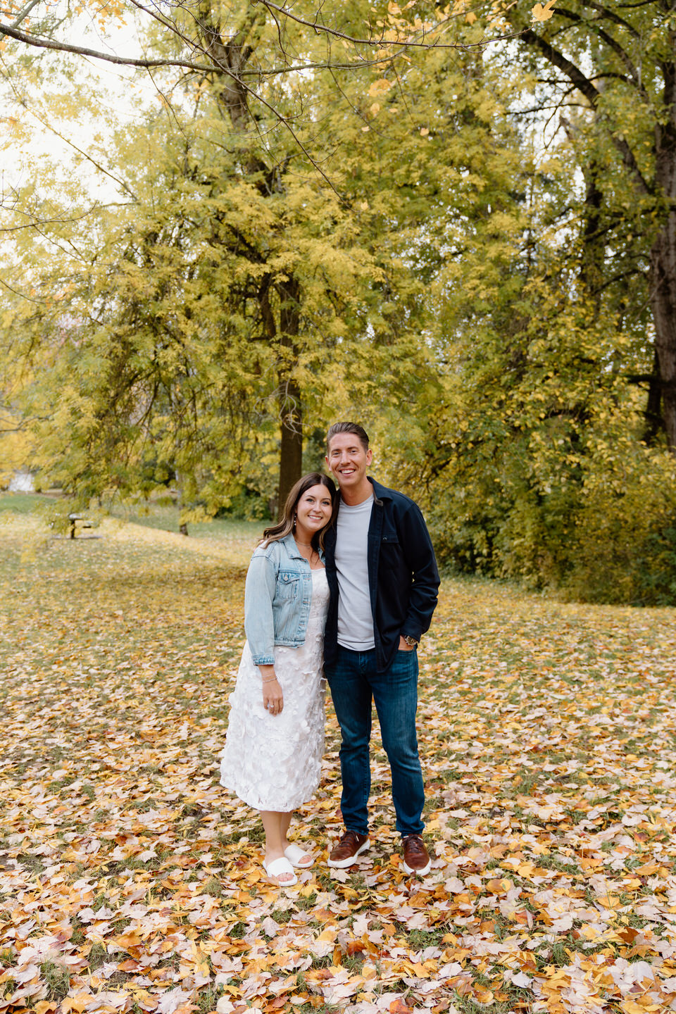 Couple smiles at the camera with yellow trees in the back and leaves all over the ground at Cathedral Park.