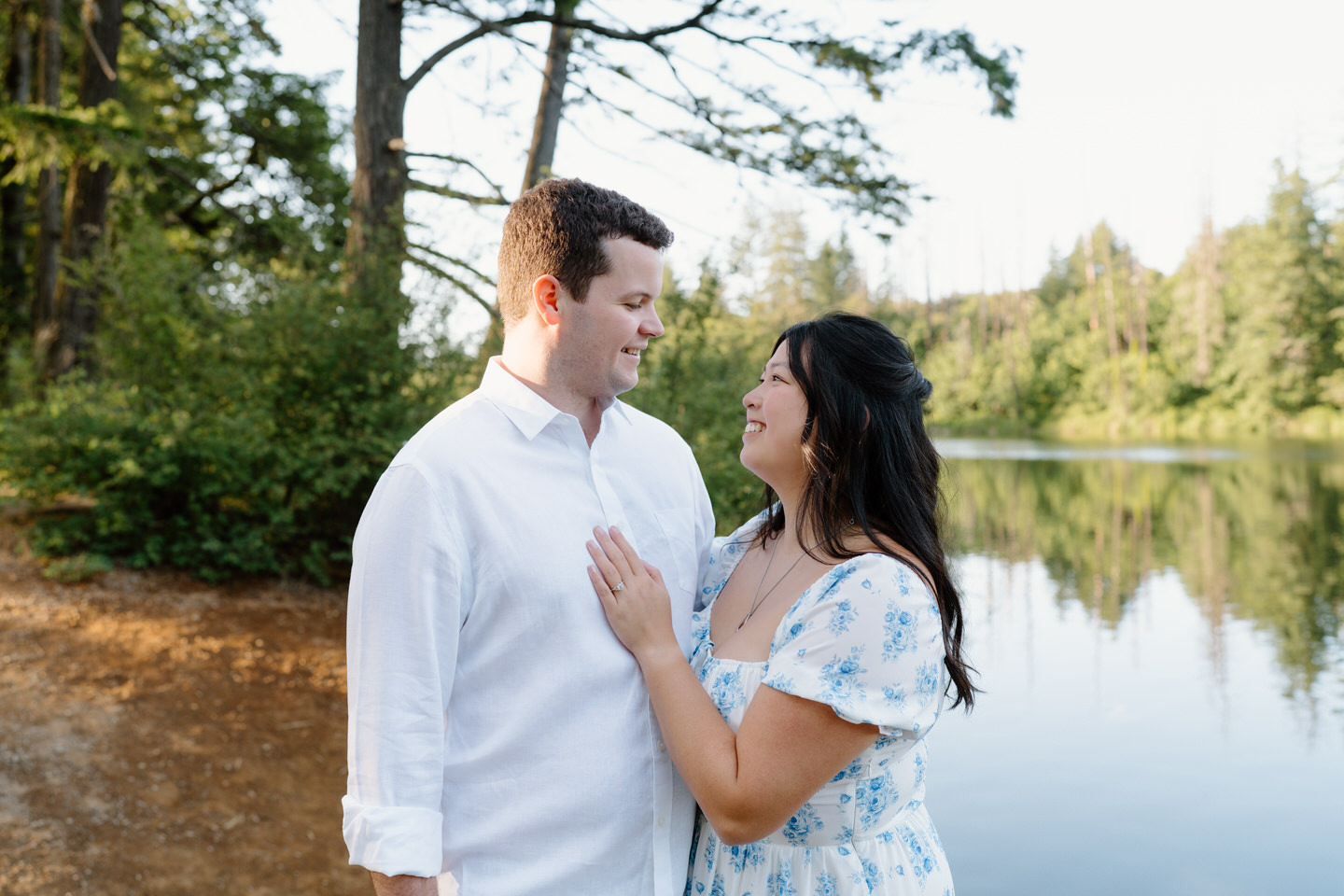 Standing by the lake, they face each other lovingly with trees mirrored in the water at their Vancouver, WA Engagement shoot.
