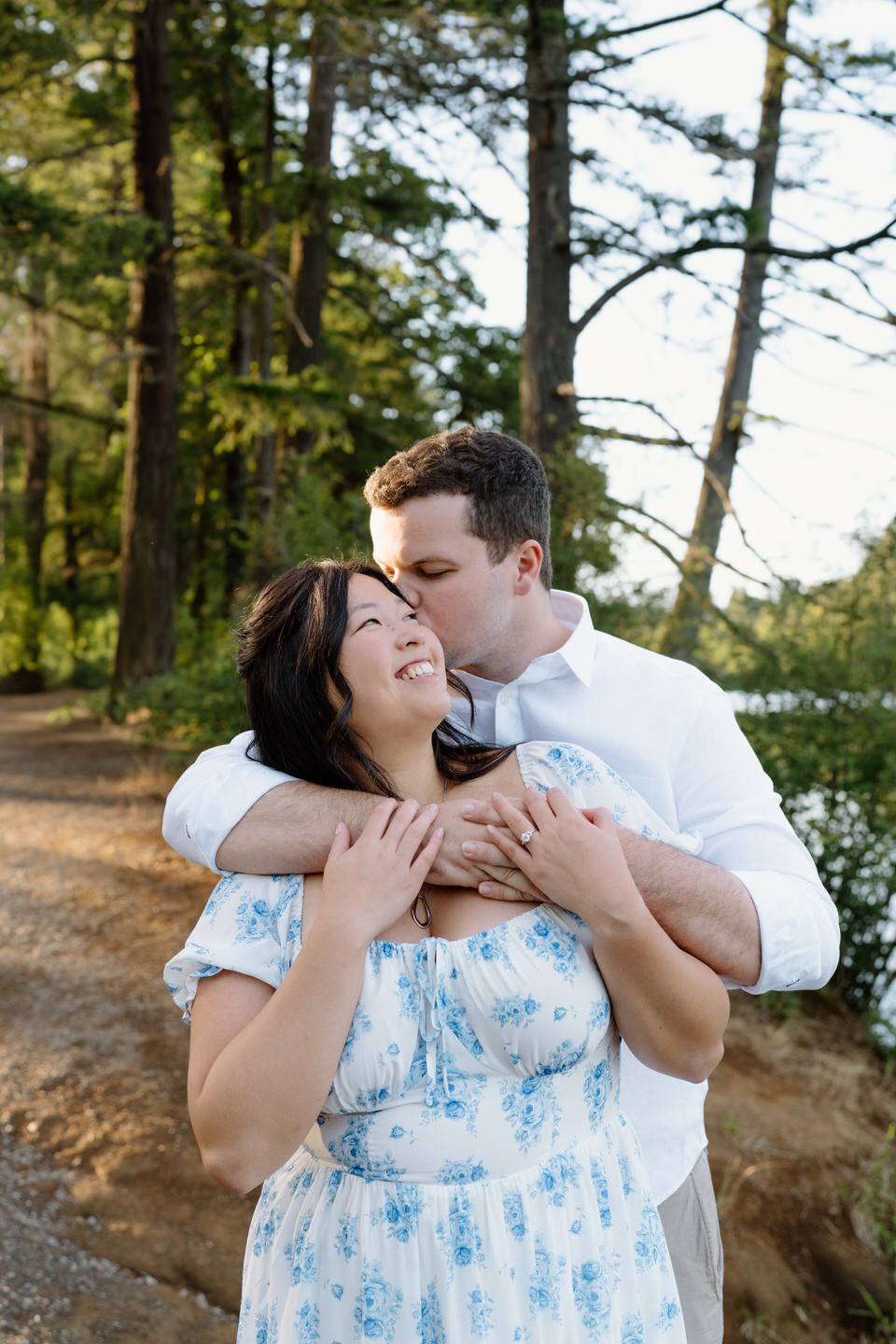He hugs her from behind near the water’s edge as she smiles, sunlight reflecting off the lake during their engagement session.