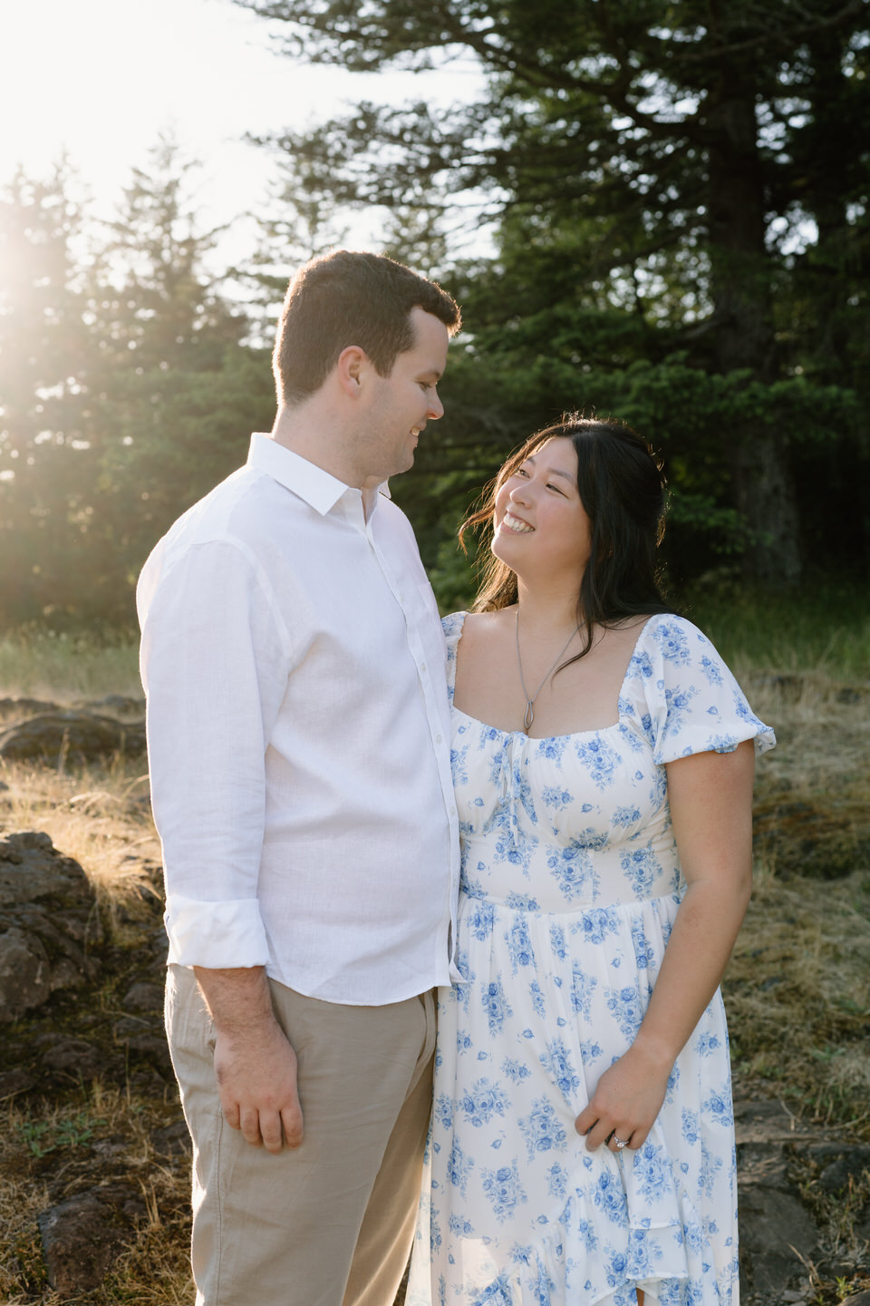 The couple stands close, smiling at each other with warm backlight and trees framing this Vancouver, WA Engagement scene.