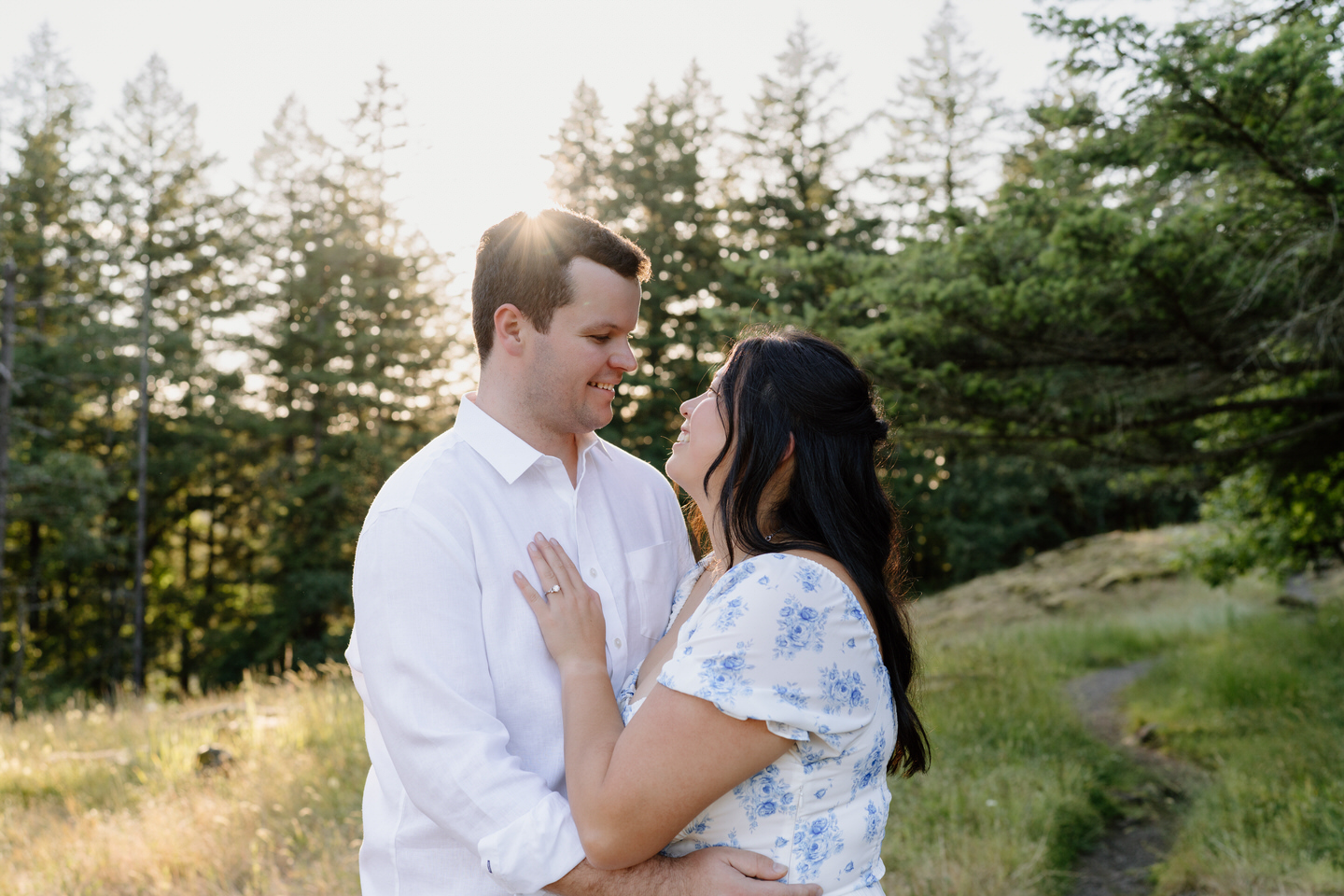 They pause mid-laugh, hands linked, framed by golden grass and evergreens on a quiet hillside.