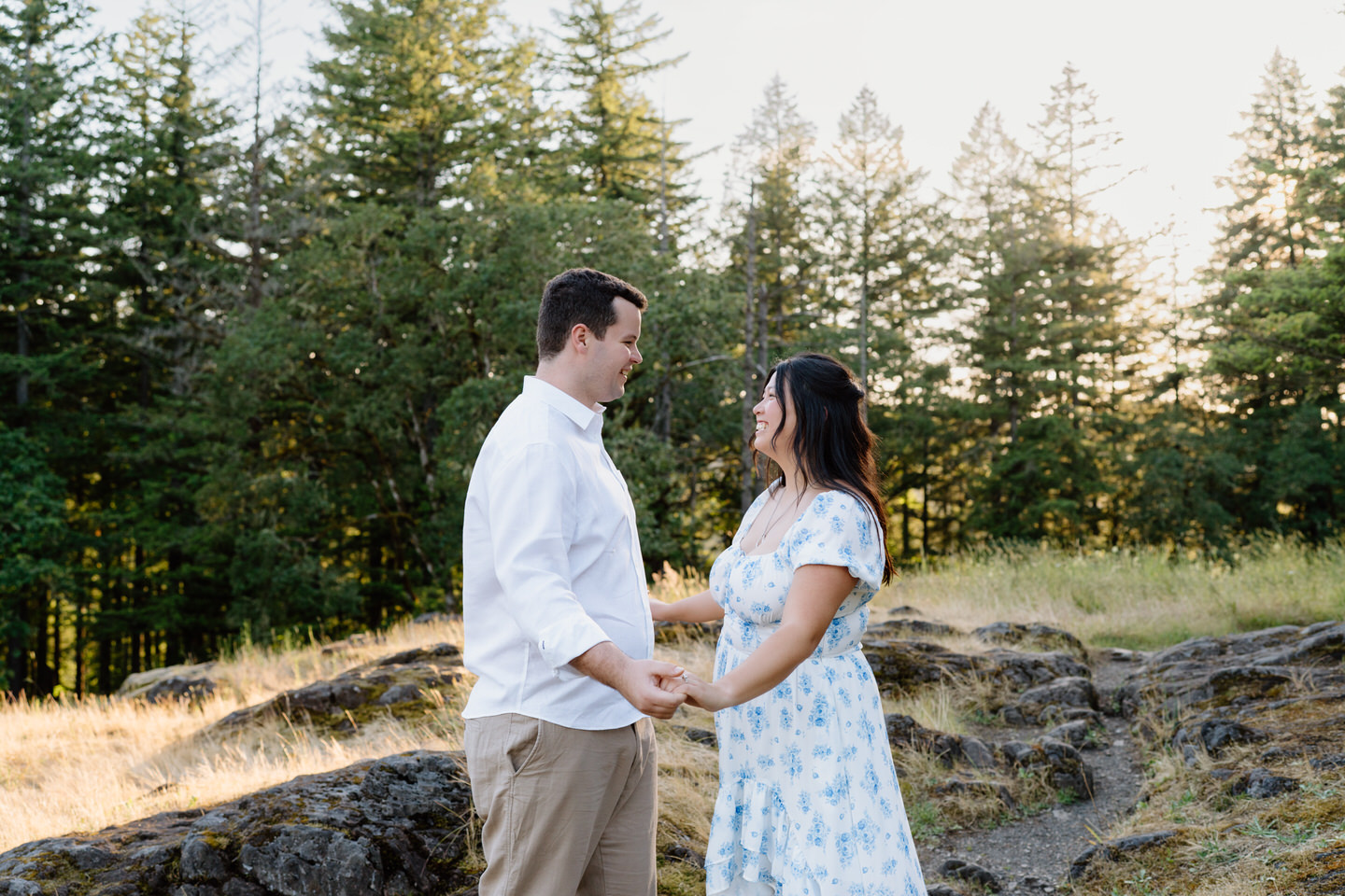 Standing hand in hand near rocky terrain, the couple gazes at each other as her dress moves in the breeze.