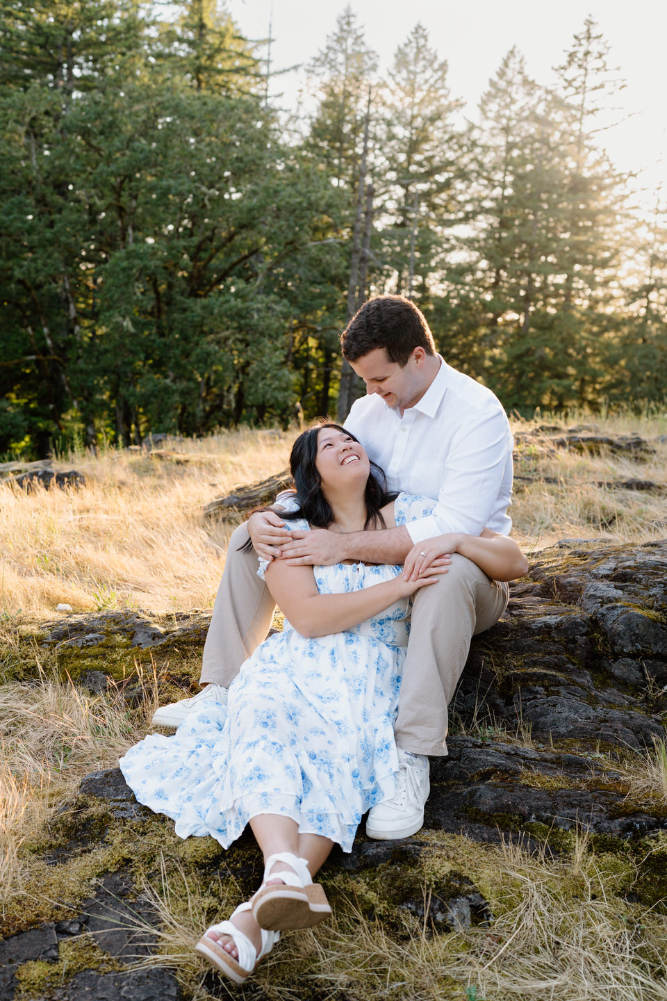 She leans back into him while seated on rocks, both smiling gently during a peaceful Vancouver, WA Engagement moment.