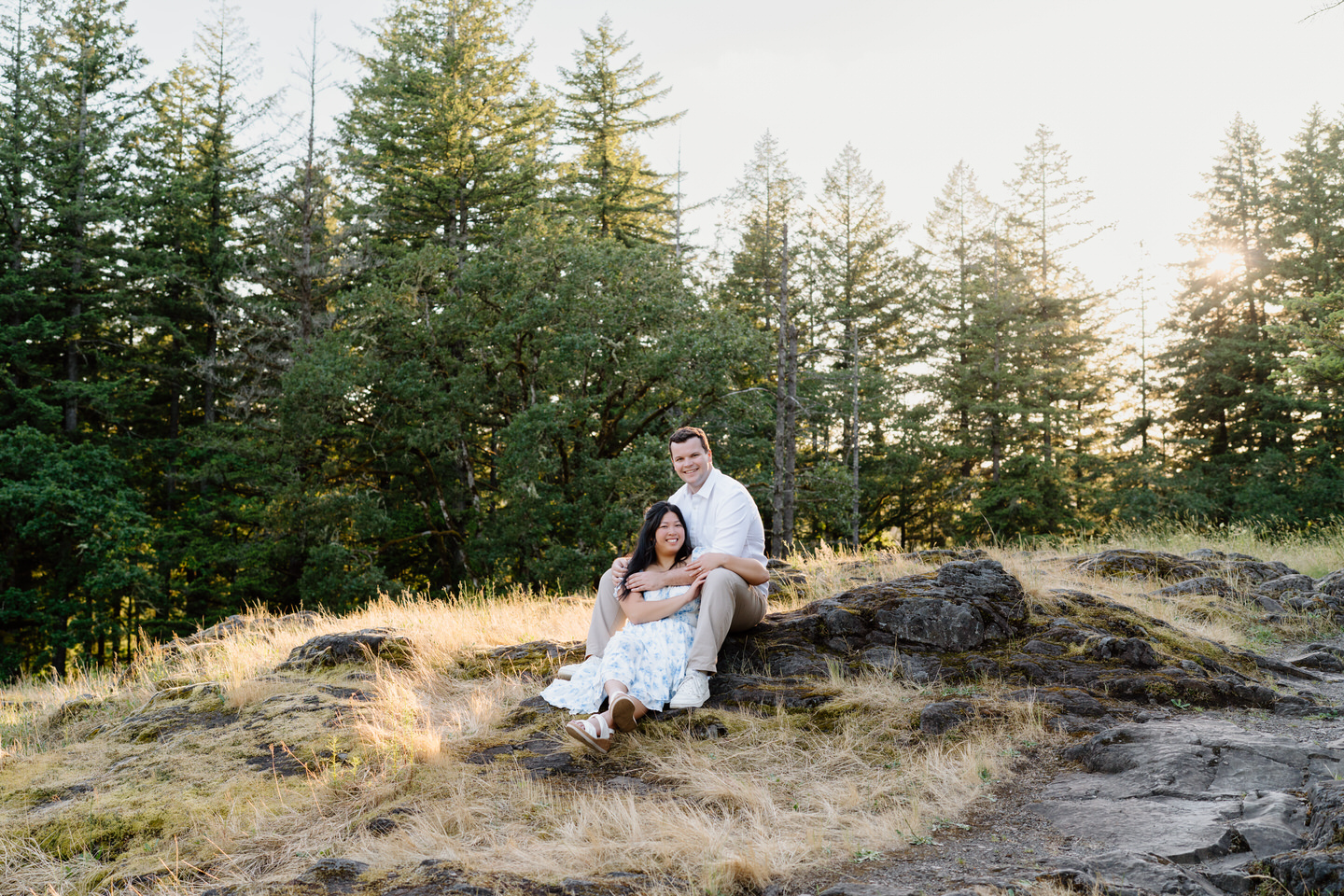 The couple relaxes together on a rocky overlook, nestled close with evergreen trees and open sky behind them.