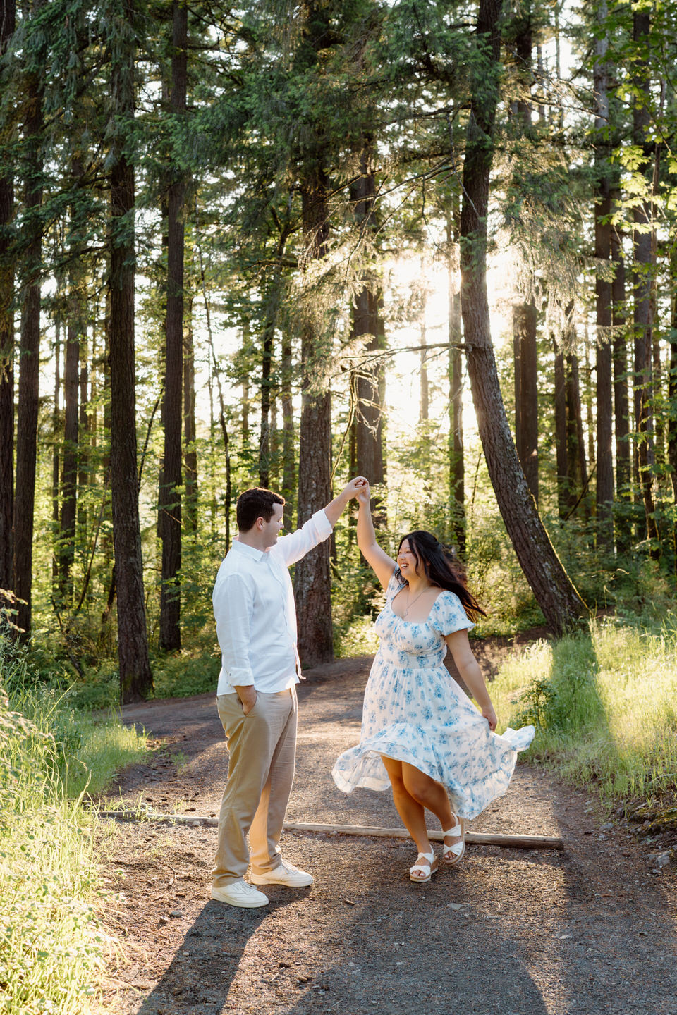 He twirls her playfully on a forest path, her blue floral dress flowing during a joyful Vancouver, WA Engagement shoot.