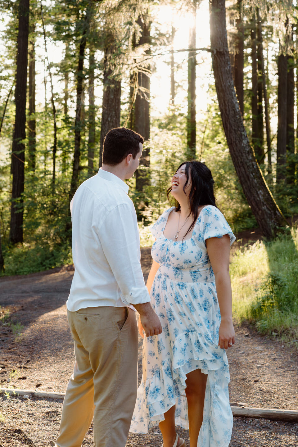 The couple faces each other on a wooded trail, sharing a quiet smile beneath towering trees and golden light.