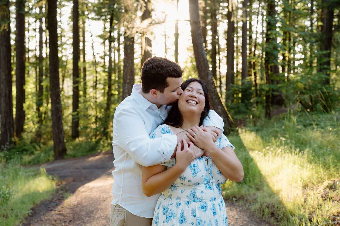 He wraps his arms around her from behind while she laughs, sunlight filtering through the trees in this romantic Vancouver, WA Engagement moment.