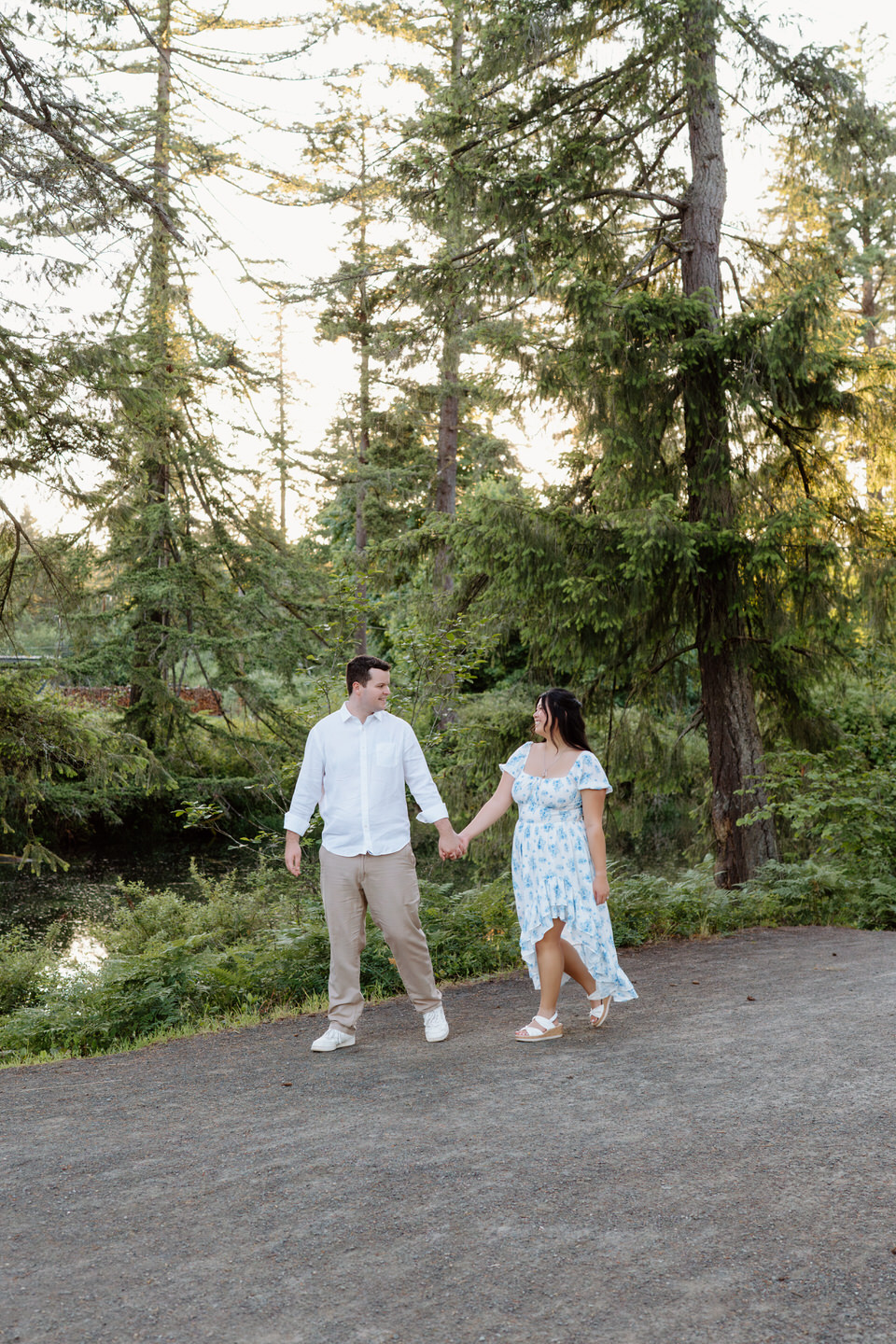 Couple walks hand in hand along a forest path, surrounded by tall evergreens and soft evening light during a Vancouver, WA Engagement session.