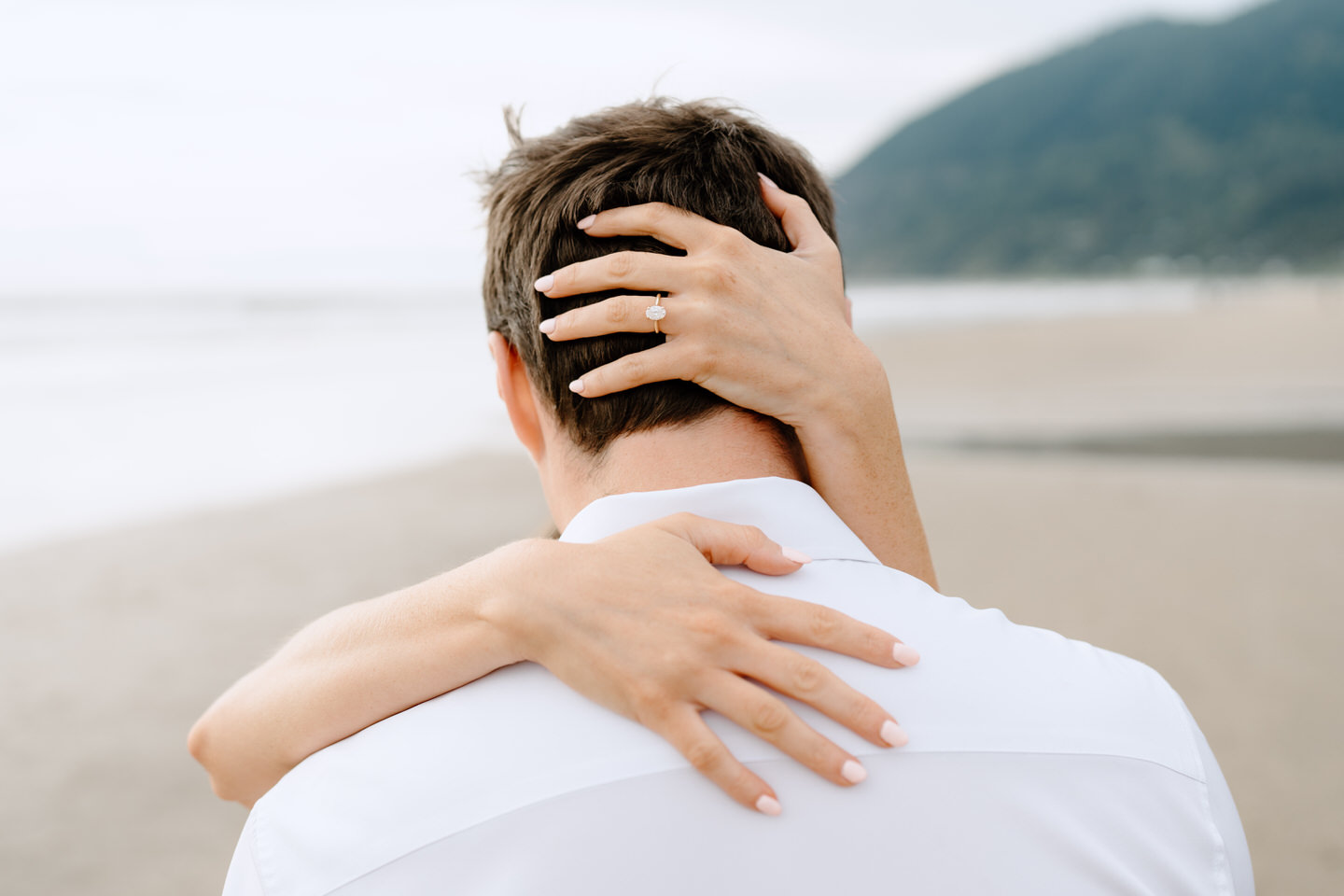 Woman wraps her hands around her fiancé's head, showing off her oval engagement ring.