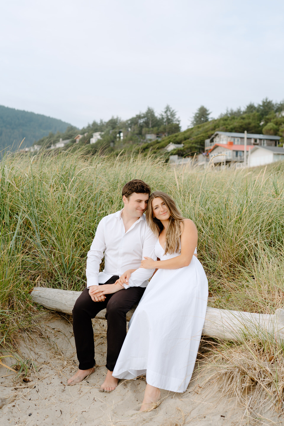 Couple sits on a log in the beach grass during Manzanita engagement photos.