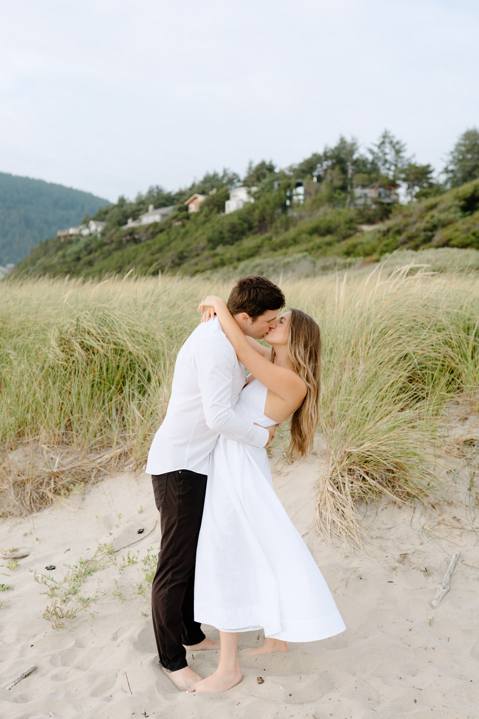 Couple wraps their arms around each other, leaning in for a kiss with her dress flowing in the ocean wind.