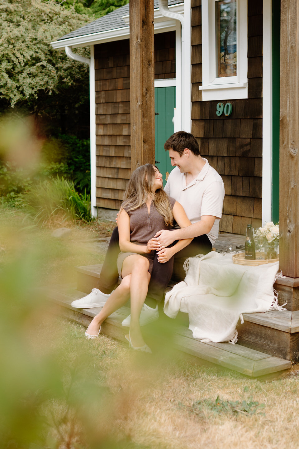 Woman sits on stairs in front of her fiancé at their beach house for Manzanita engagement photos.