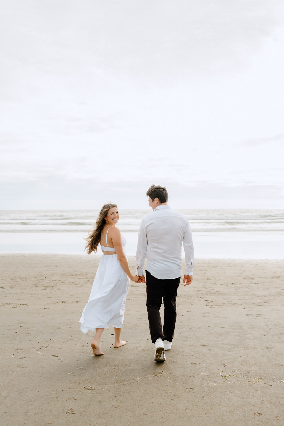 Couple walks away, towards the ocean, with the woman looking back over her shoulder. 