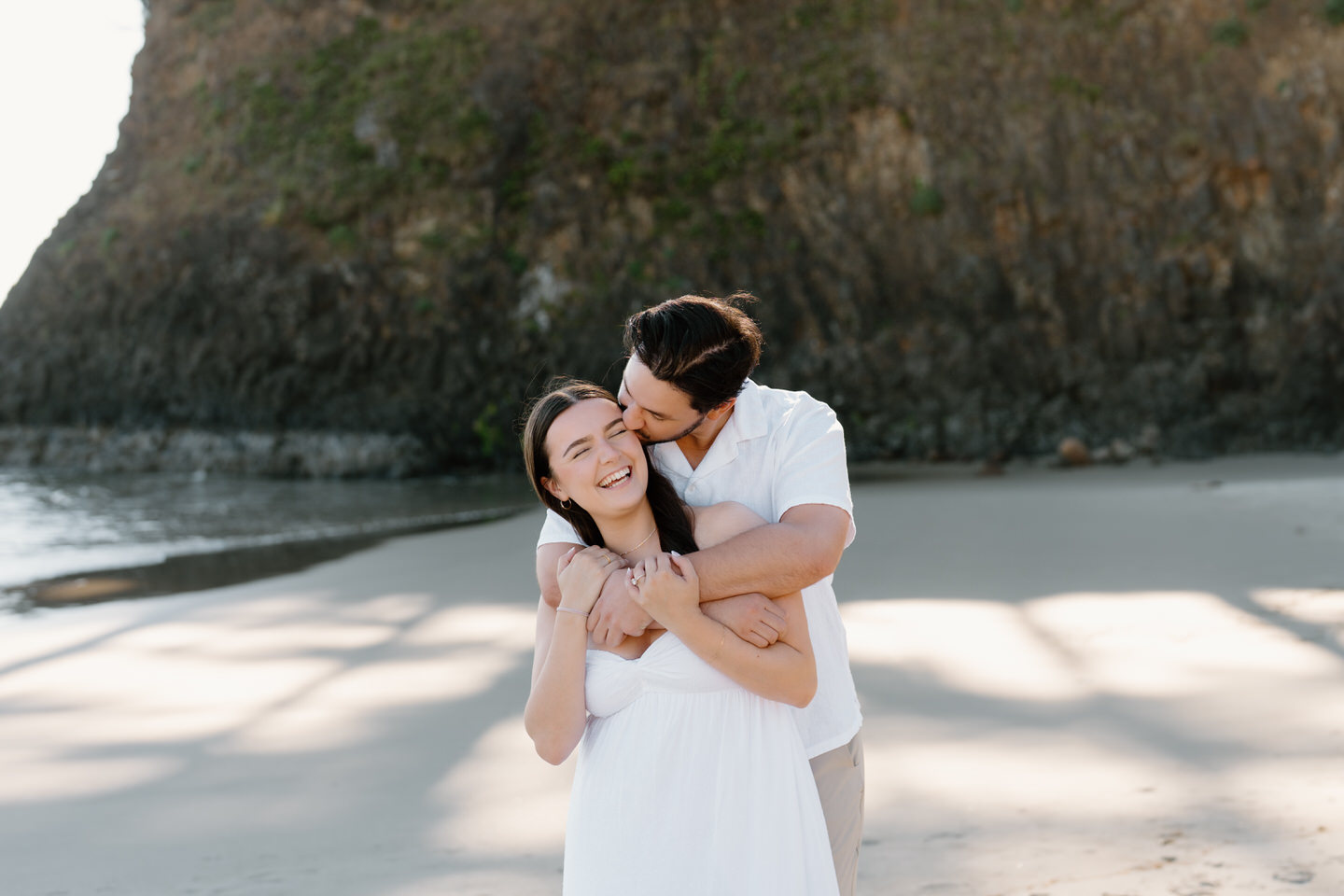 Man wraps his arms over his fiancee's shoulders, giving her a kiss on the cheek.