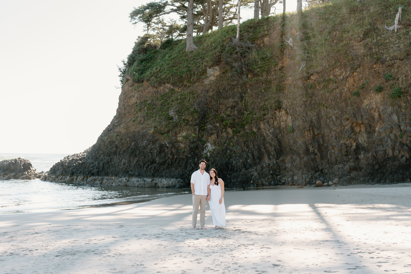 Couple looks at the camera, posing in front of the Oregon coast cliffs with rays of sun shining through.