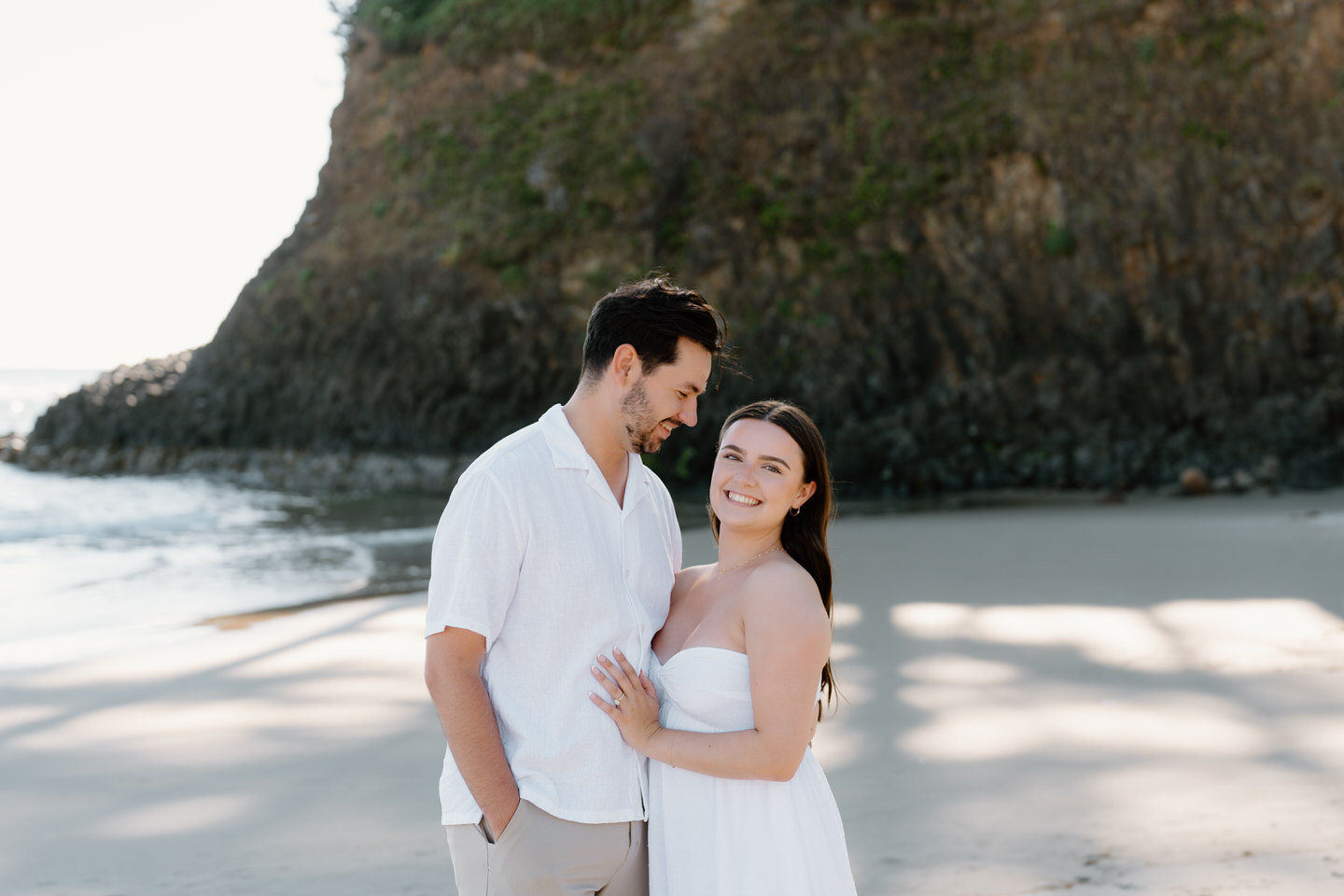 Woman looks at camera while man looks at her, both in white attire for their engagement photos.
