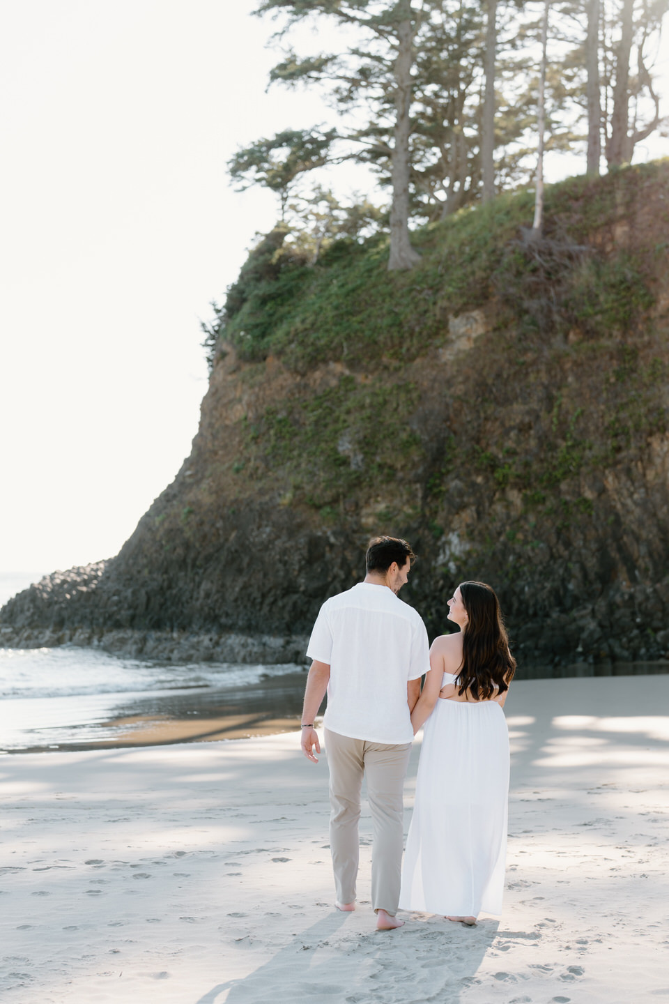 Couple walks away towards the Oregon Coast cliffs, holding hands and barefoot in the sand.