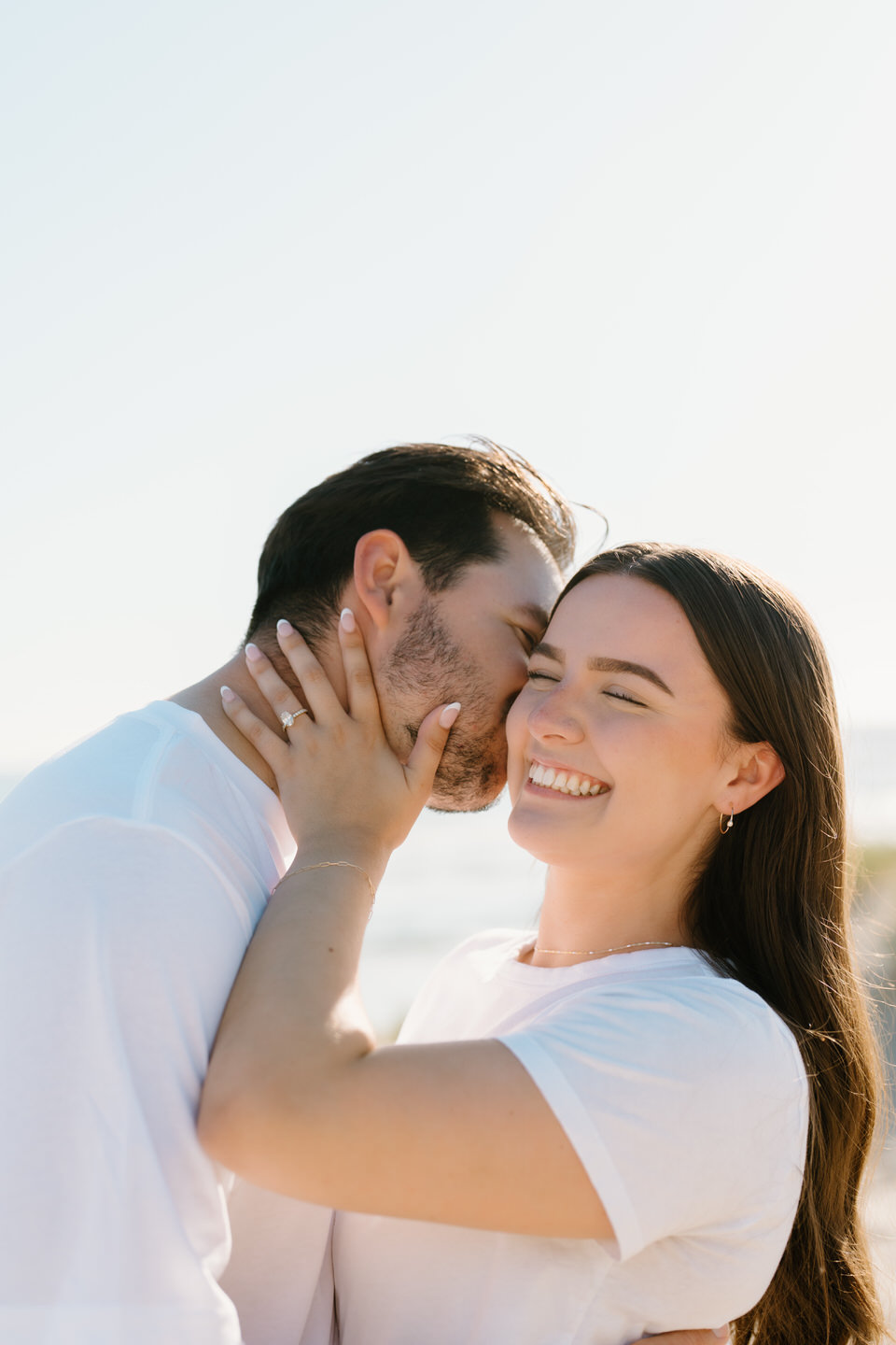 Man kisses woman on the cheek while she holds his face, showing off her engagement ring.