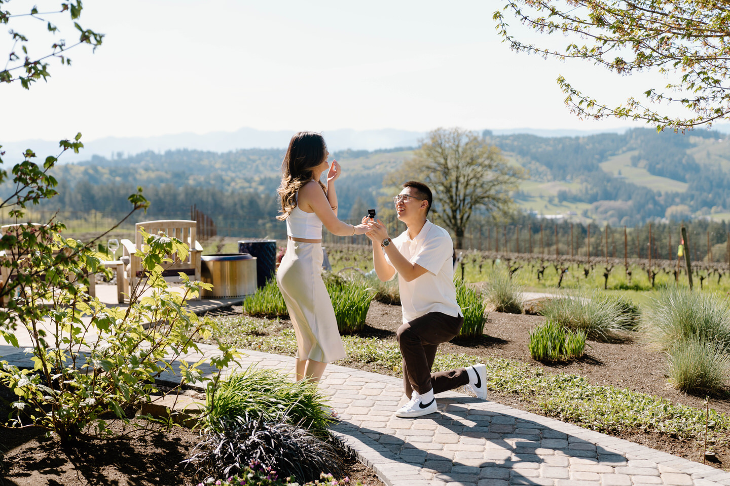 Surprise proposal scene at Eminent Domaine winery as partner kneels on stone path surrounded by vineyards.