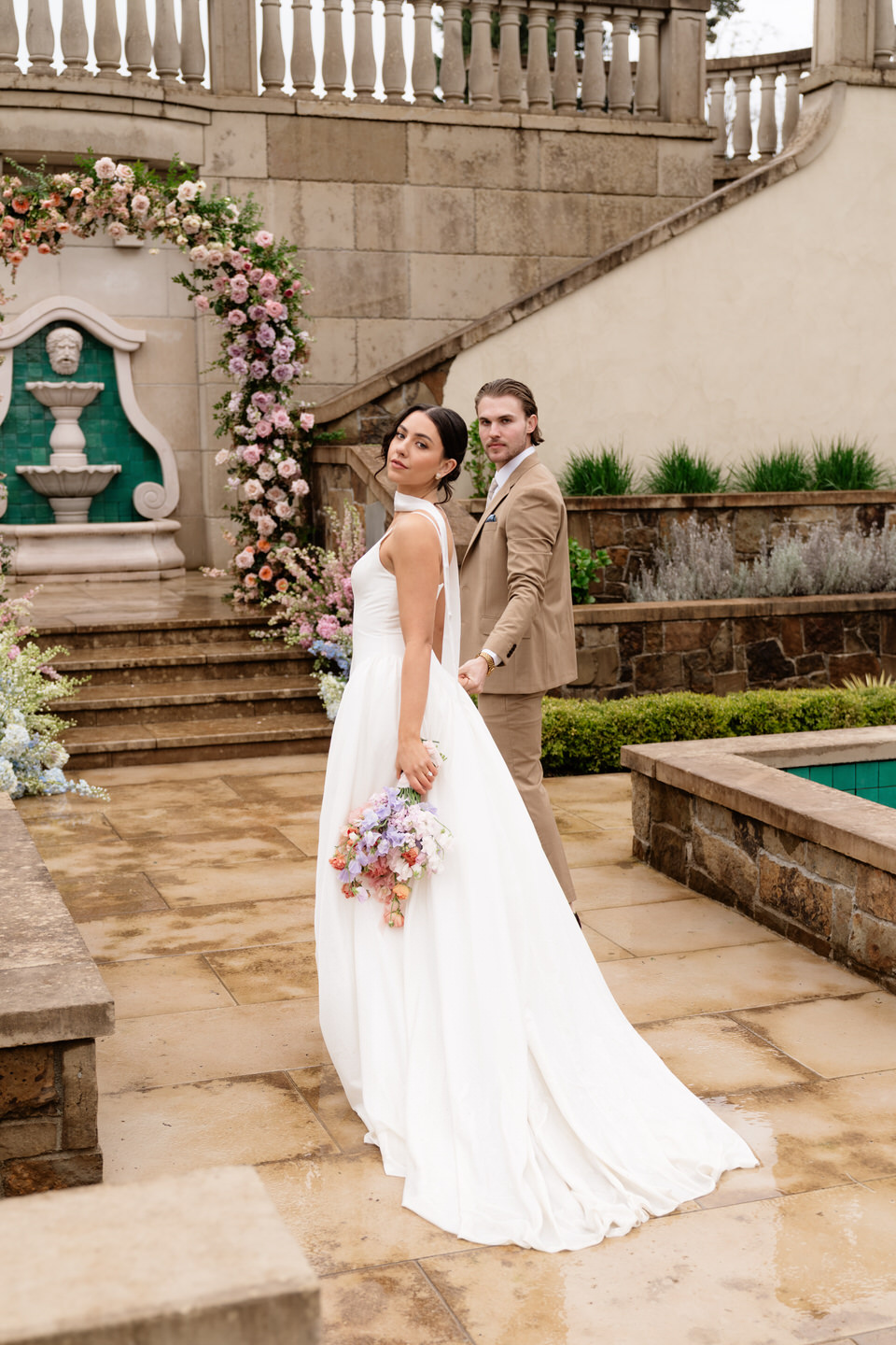 The bride turns gracefully as the groom looks on, captured in a refined courtyard with stone details.