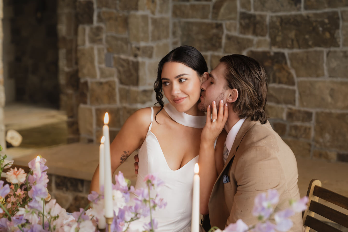 Groom whispers into the bride’s ear while she smiles, framed by candles and spring blooms on the table.