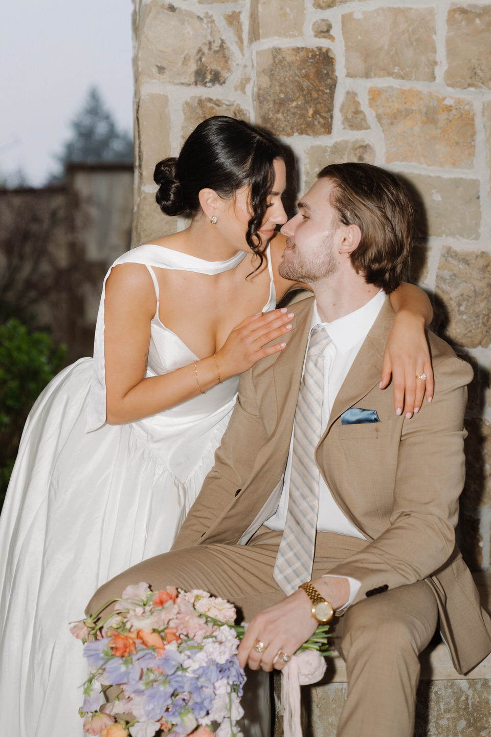 Bride kisses the groom’s cheek while seated together, capturing an intimate moment against warm stone textures.