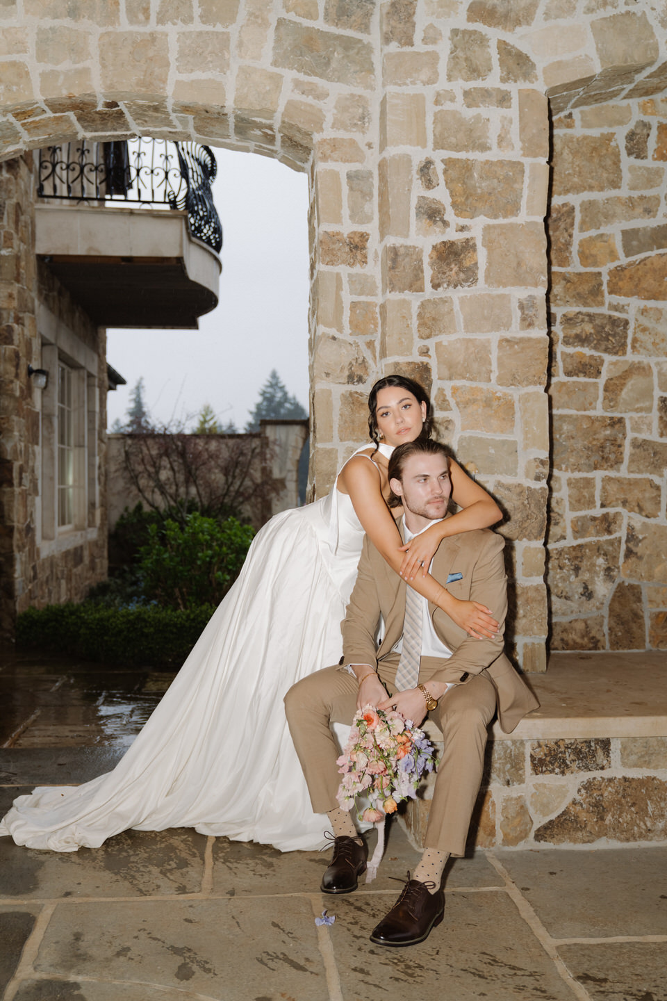The groom sits as the bride leans in affectionately, her veil draping across stone walls at chateau de lis.