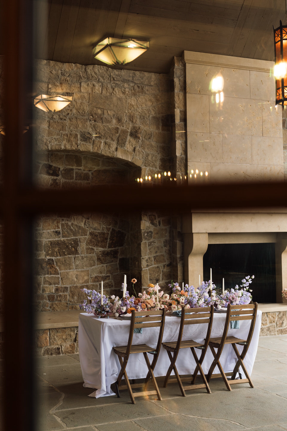 Reception table setup at Chateau de Lis with long farmhouse tables, pastel florals, and candlelight ambiance, shot through a window.