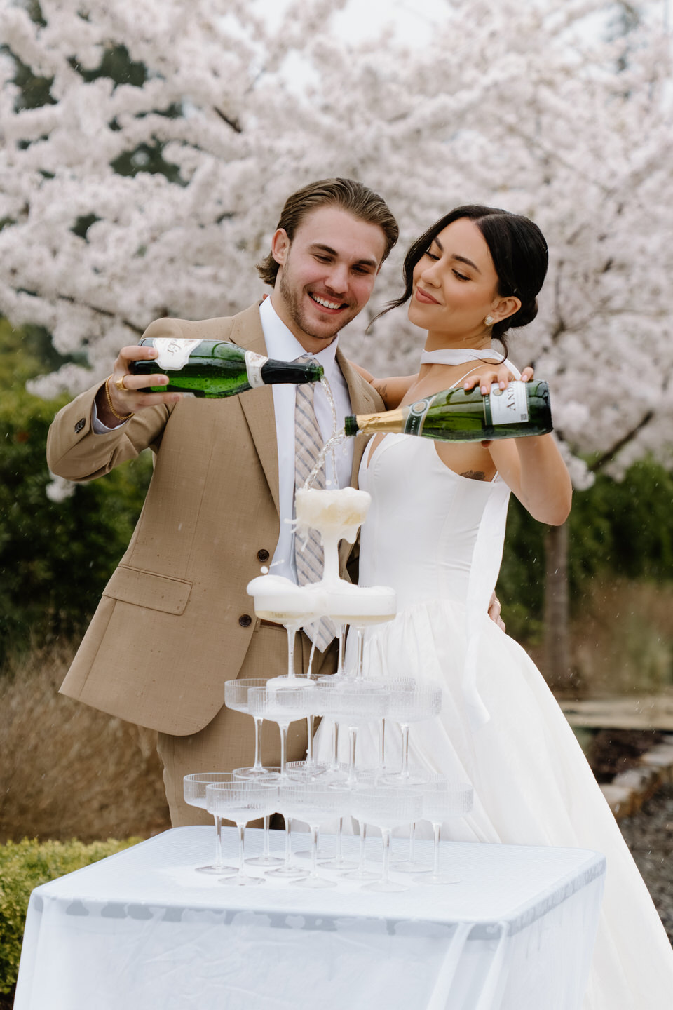 Couple pouring champagne out of two bottles over a tower of champagne glasses.