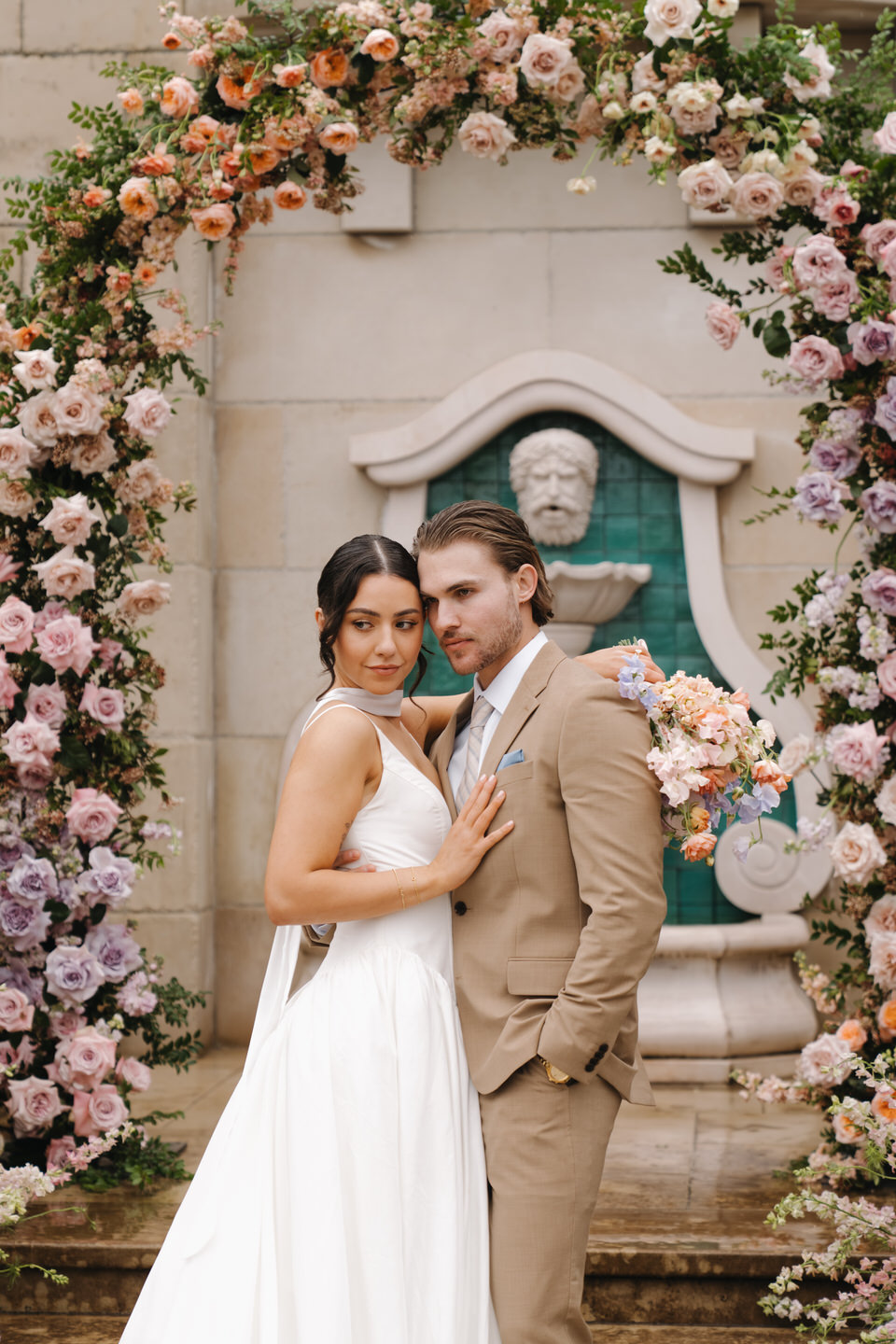 Bride and groom stand centered beneath the arch, posed symmetrically with soft pastel florals at chateau de lis.