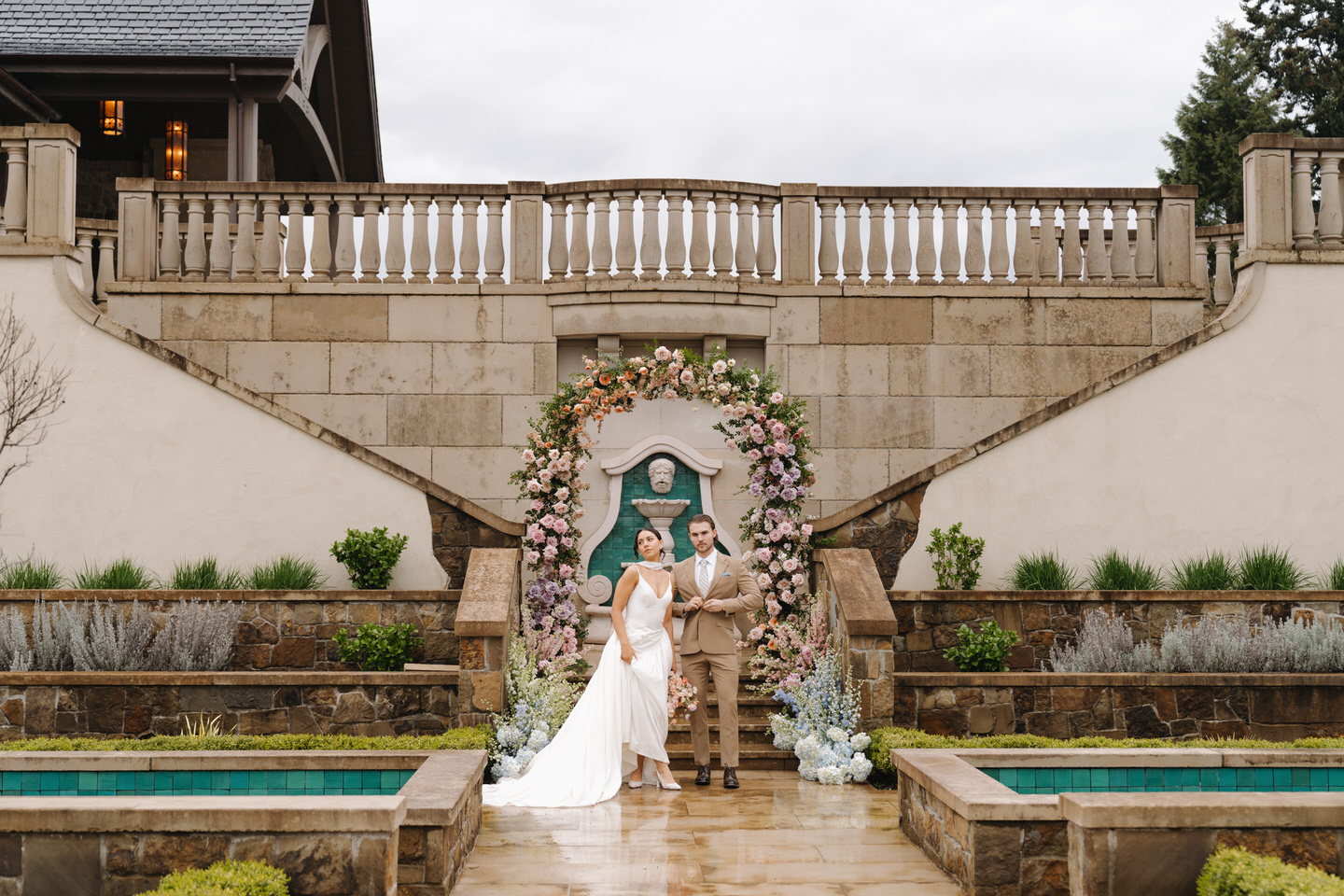 Romantic portrait of the couple framed by lush florals and stone architecture in a European-style courtyard.