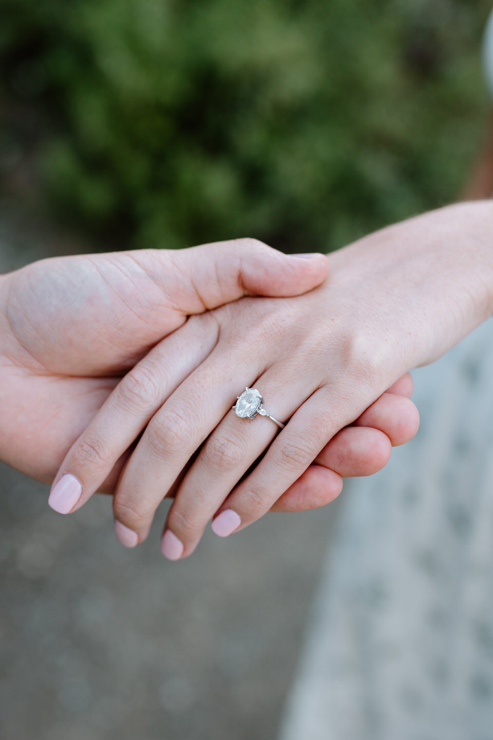 Close-up of hands showing oval engagement ring with natural greenery background.